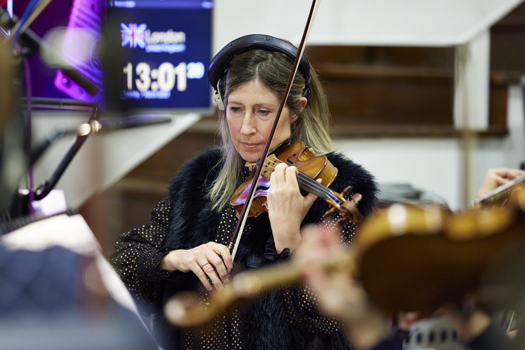 Une femme joue du violon lors d'une séance d'enregistrement ou de répétition, portant des écouteurs et concentrée sur sa musique, avec un écran affichant l'heure derrière elle.