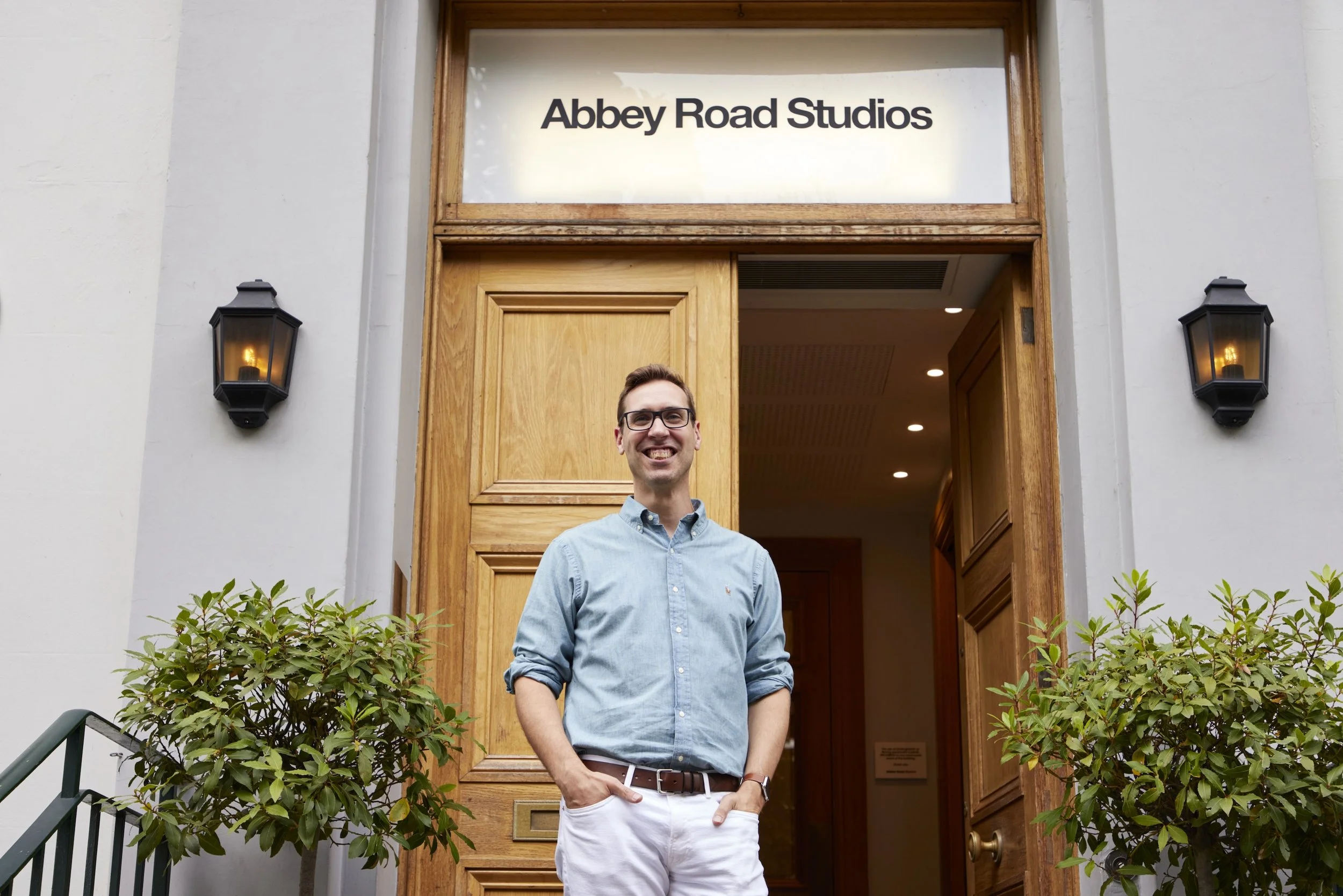 Un homme souriant en chemise bleue et pantalons blancs se tient devant l'entrée de Abbey Road Studios, entouré de buissons verts, sous la lumière de deux appliques murales.