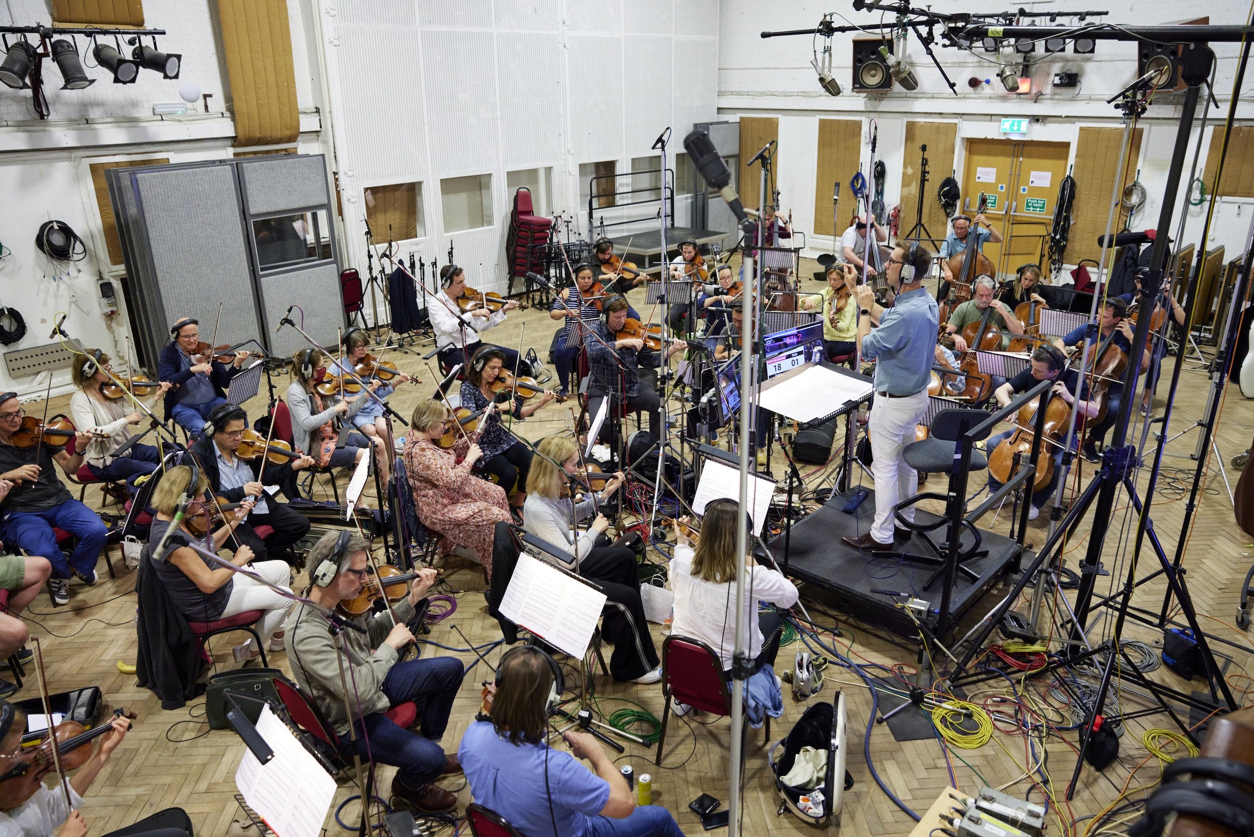 Un grand ensemble d'orchestre symphonique en train de jouer, avec un chef d'orchestre sur une plateforme, dans un studio d'enregistrement équipé de microphones et de câbles.