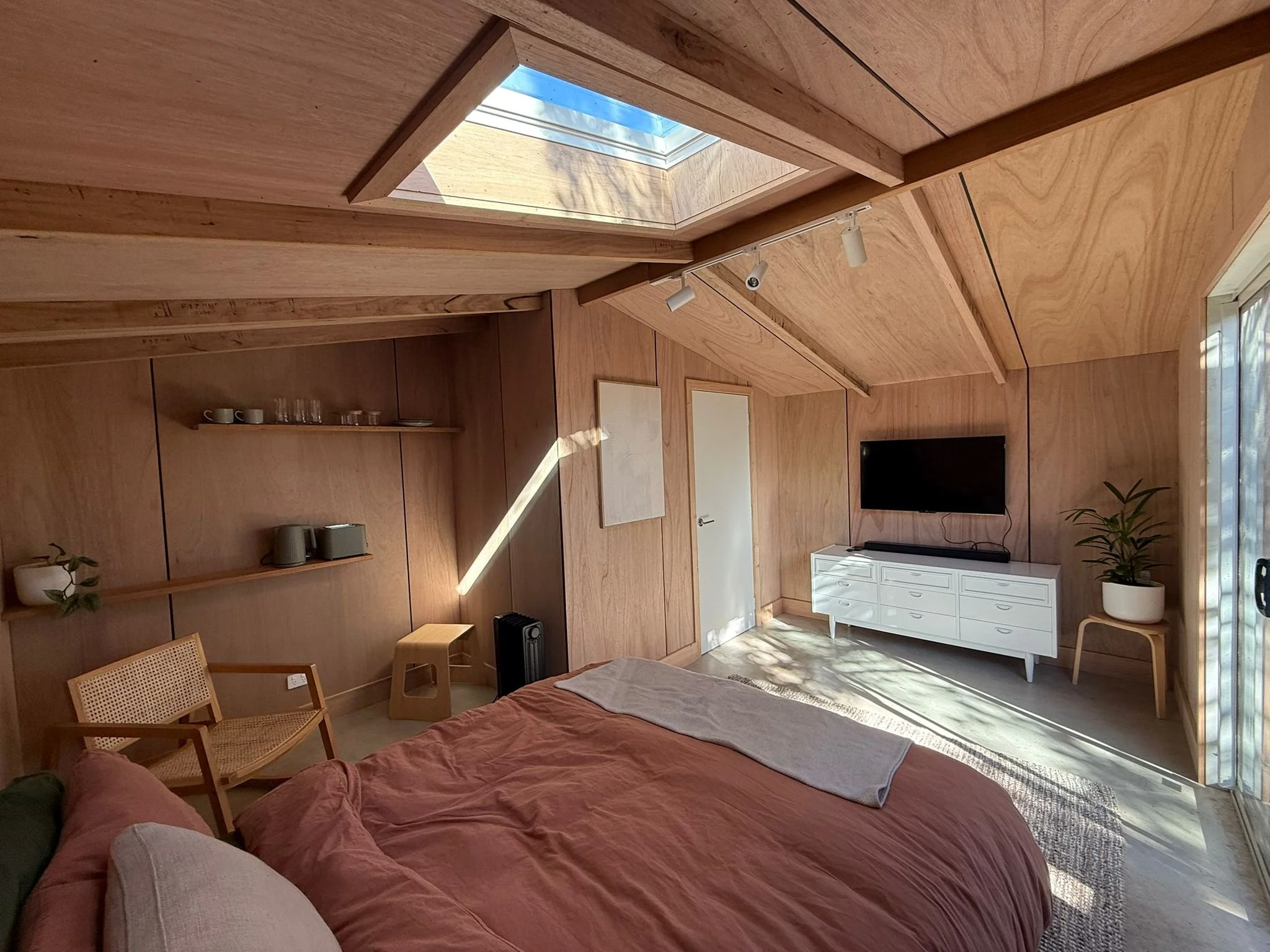 A cozy bedroom with wooden walls and a slanted ceiling with a skylight. There is a bed with pink bedding, a white dresser with a flat-screen TV above it, and potted plants. Natural sunlight illuminates the room.