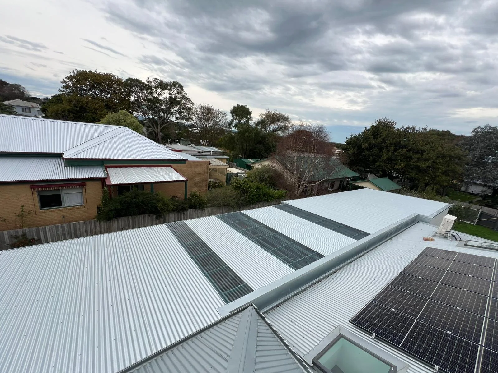 Aerial view of rooftops with solar panels and metal roofing in a residential neighborhood with trees and cloudy sky.