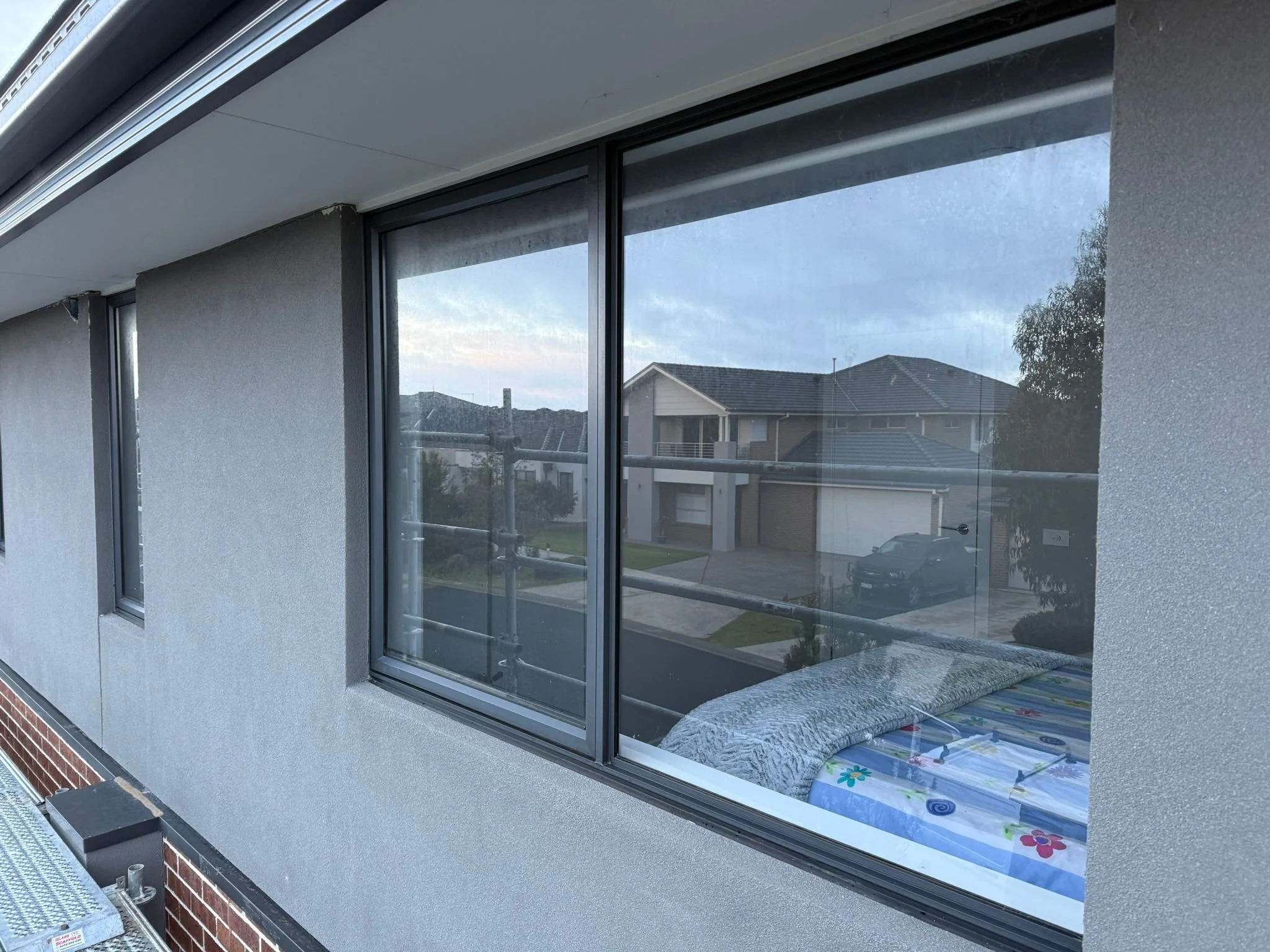 View of a house exterior through a large window, showing a bedroom with a bed and a neighborhood street with houses and parked cars.