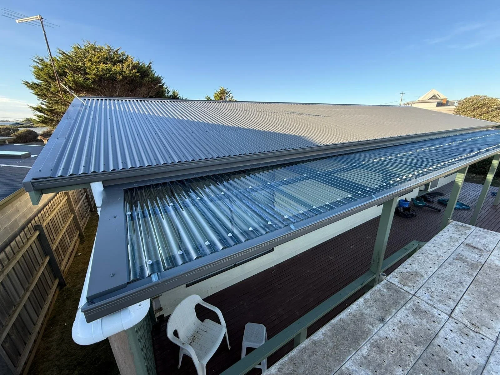 View of house with partially installed corrugated metal roof, including glass panels, slate flooring, a white chair, and a clear sky.