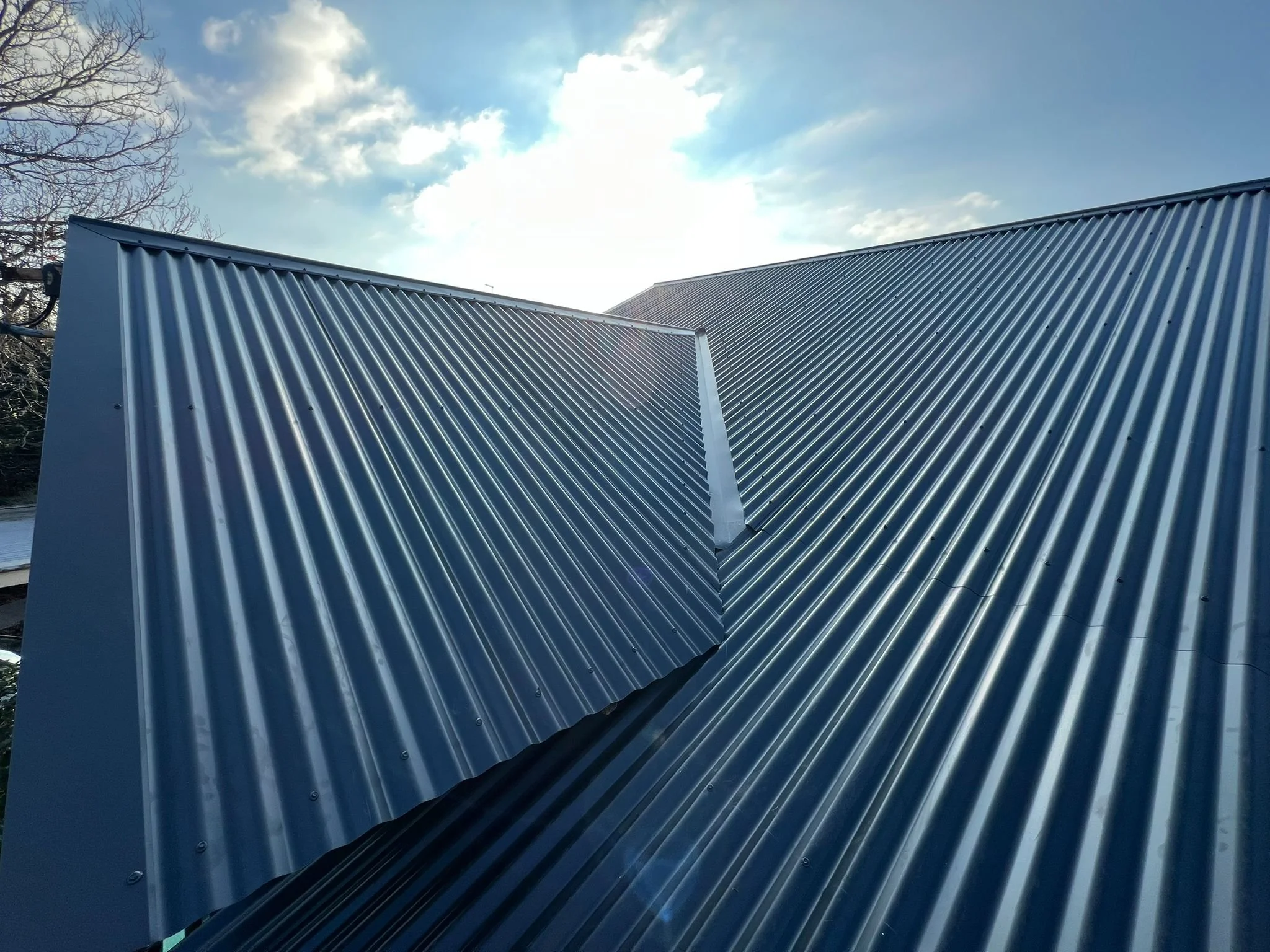 Metal roof with ridges on a building, under a partly cloudy sky with the sun shining.