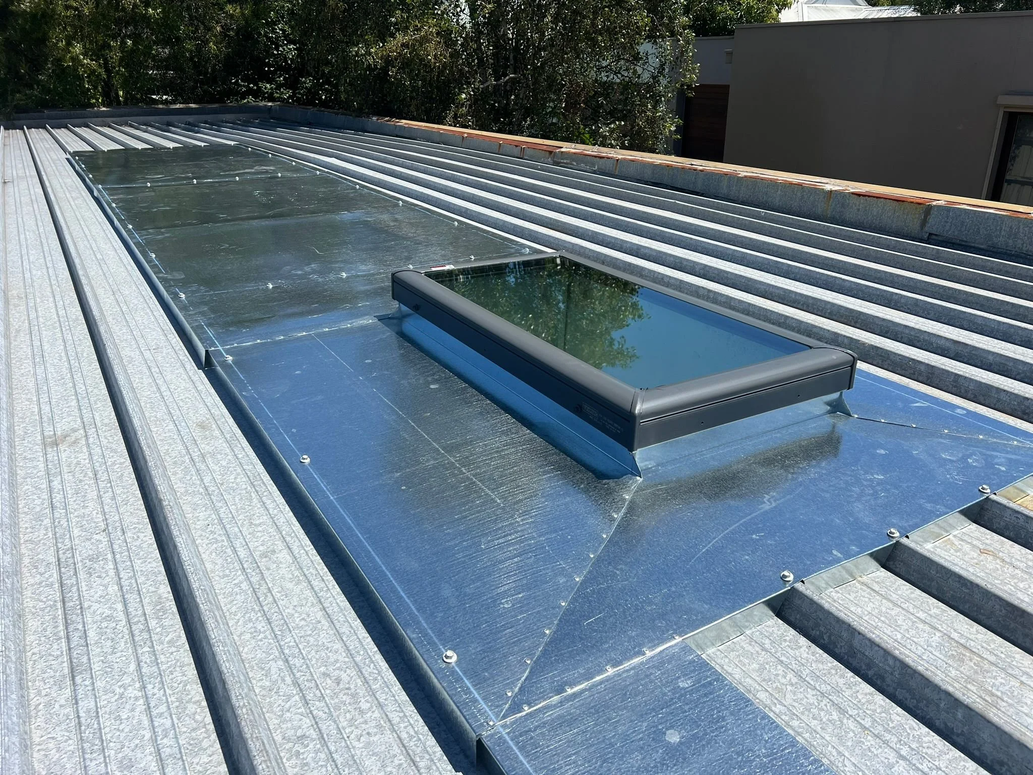 Metal roof with skylight window, surrounded by trees and neighboring buildings.