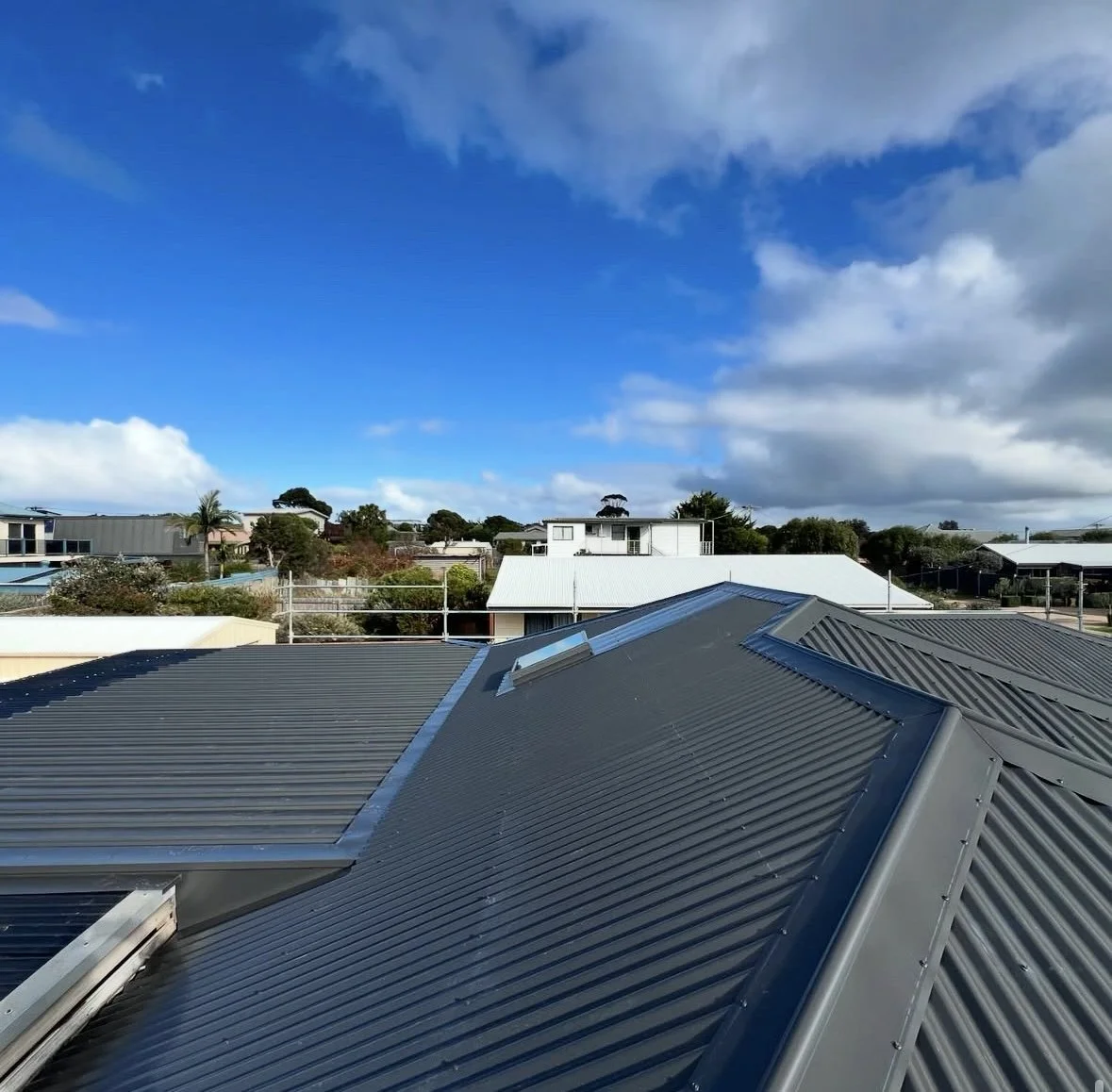 View of rooftops with metal roofing, a partly cloudy sky, and residential houses and trees in the background.