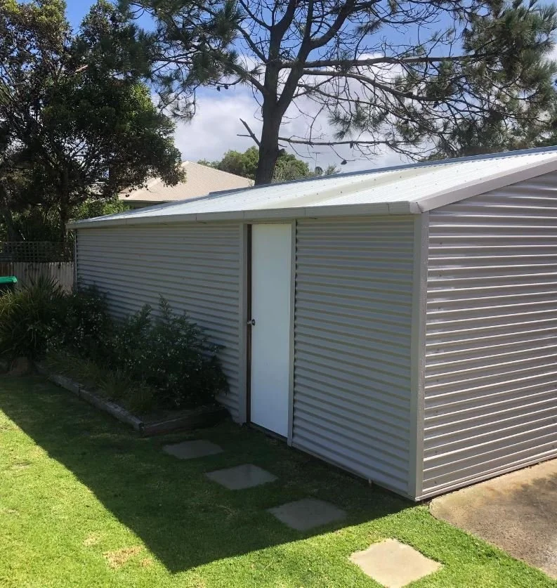 A backyard view showing a metal storage shed with a white door, surrounded by green grass, bushes, and trees, under a partly cloudy sky.