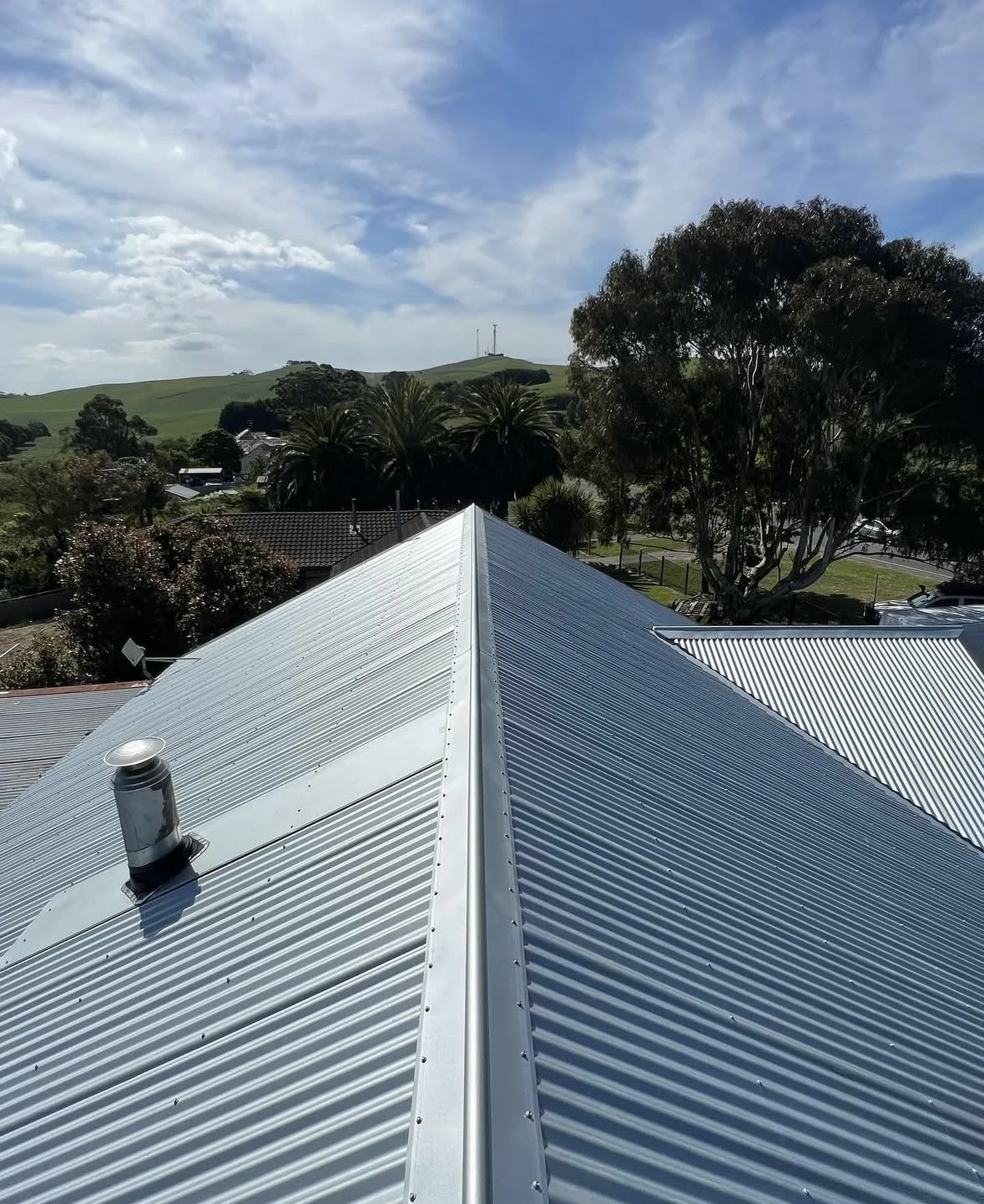 Metal roof with a vent pipe, surrounded by trees and a rural landscape under a partly cloudy sky.