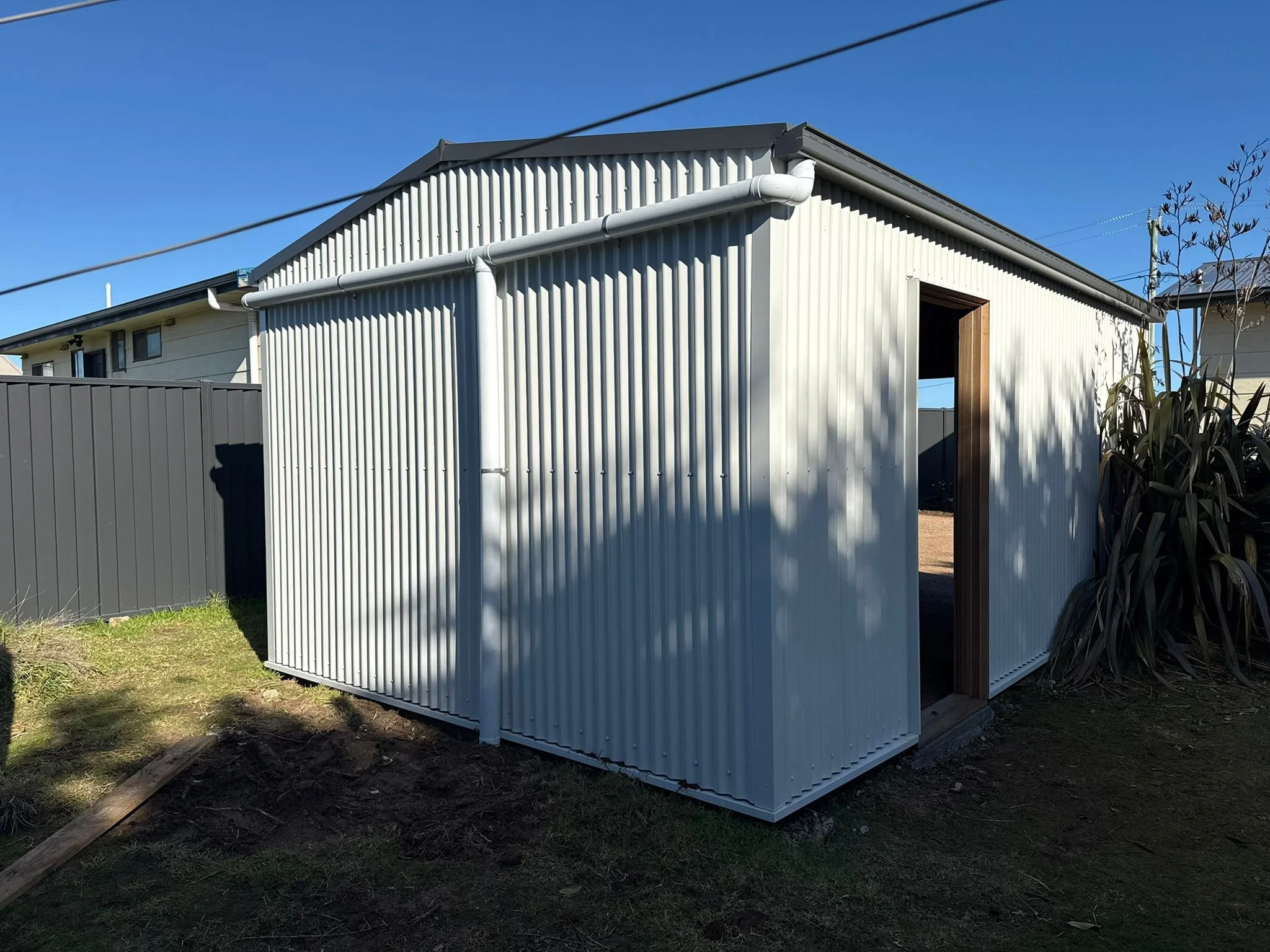 A small metal shed with a gable roof and large entryway, located in a backyard with grass and nearby plants, under a clear blue sky.