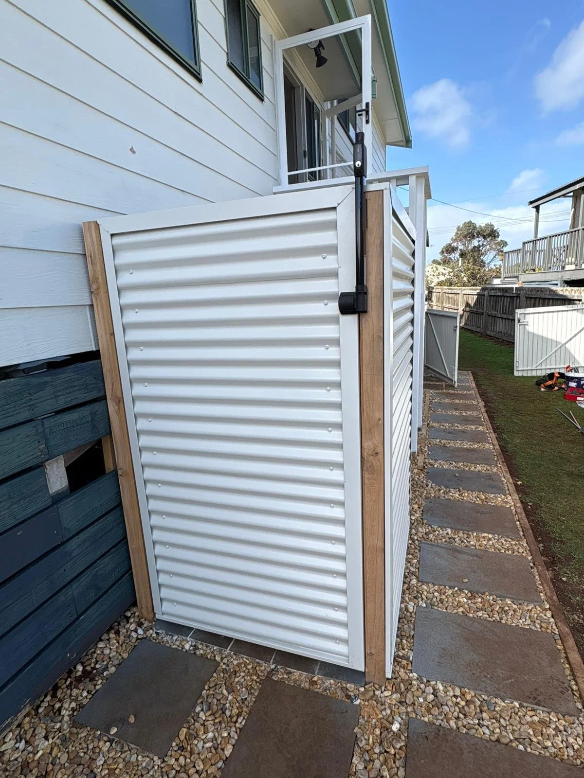 Outdoor shower enclosure with white corrugated metal panels, wooden posts, and a black mounted shower head, situated along a stone-paved pathway next to a house.