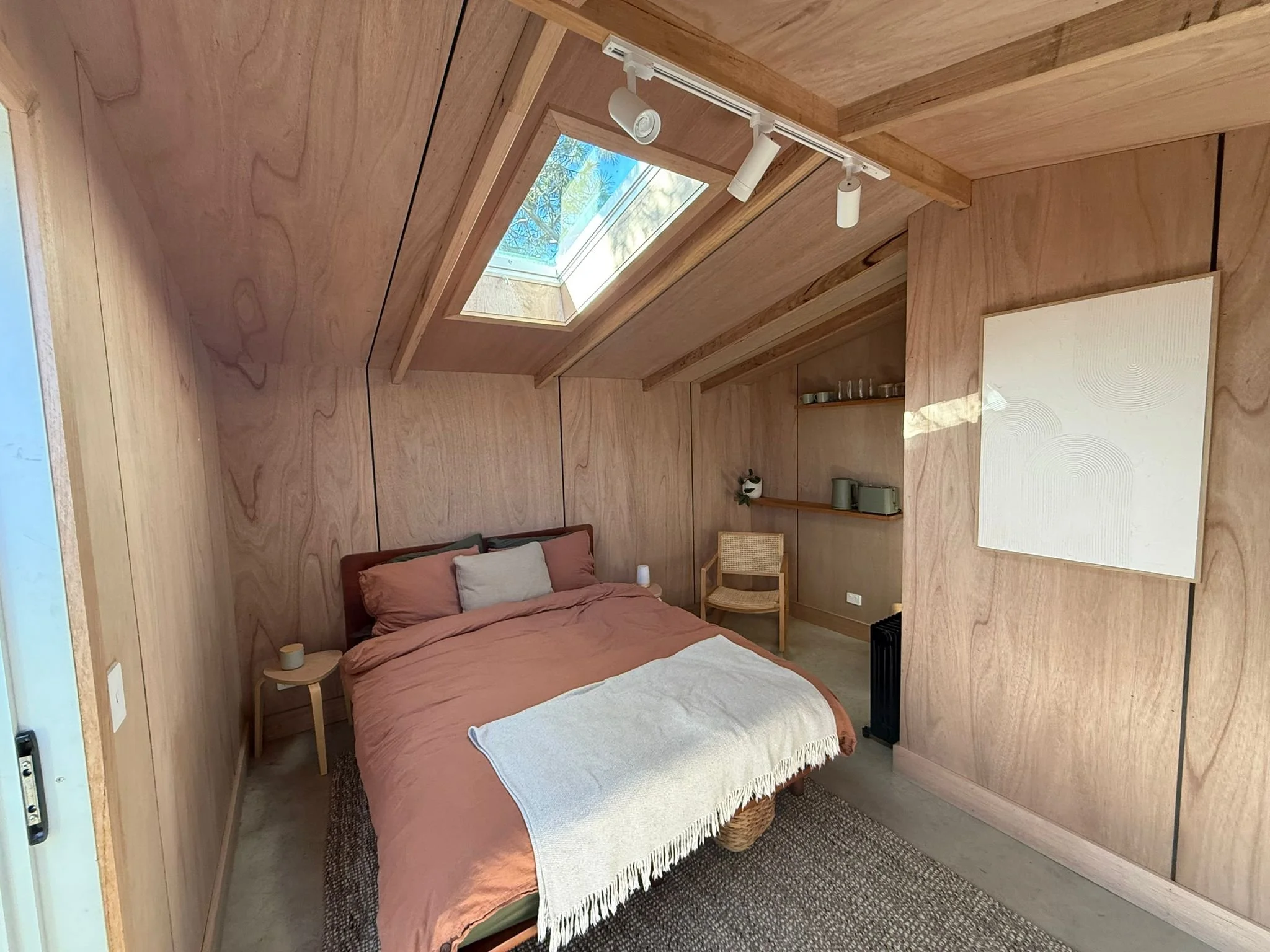 A cozy bedroom with wooden walls and ceiling, a bed with rust-colored bedding and pillows, a small side table with a candle, a wicker chair, wall shelves with decorative items, a skylight window showing blue sky and trees, and a white abstract painti