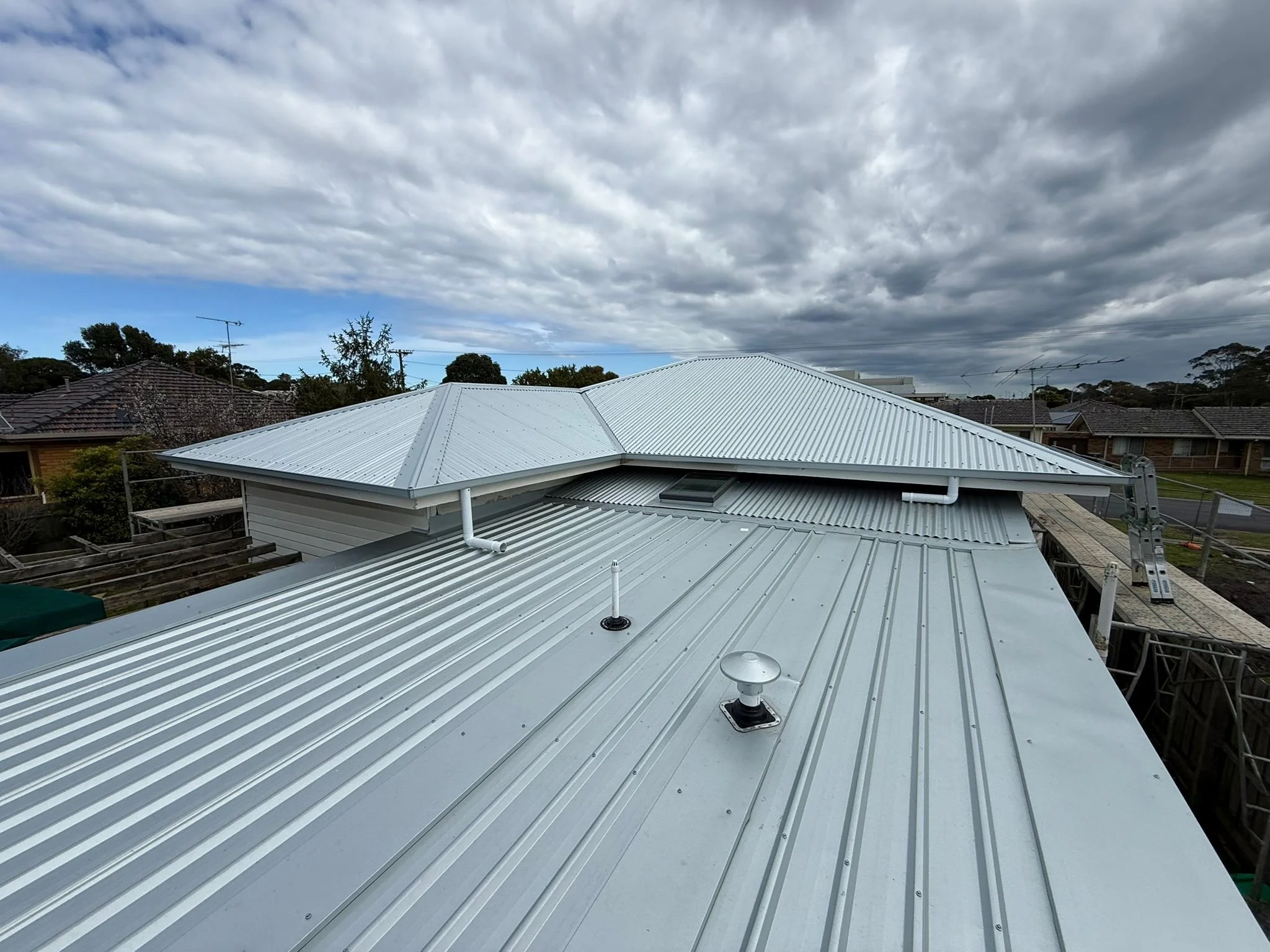 View of a metal rooftop with building structures, vent, and cloudy sky overhead.