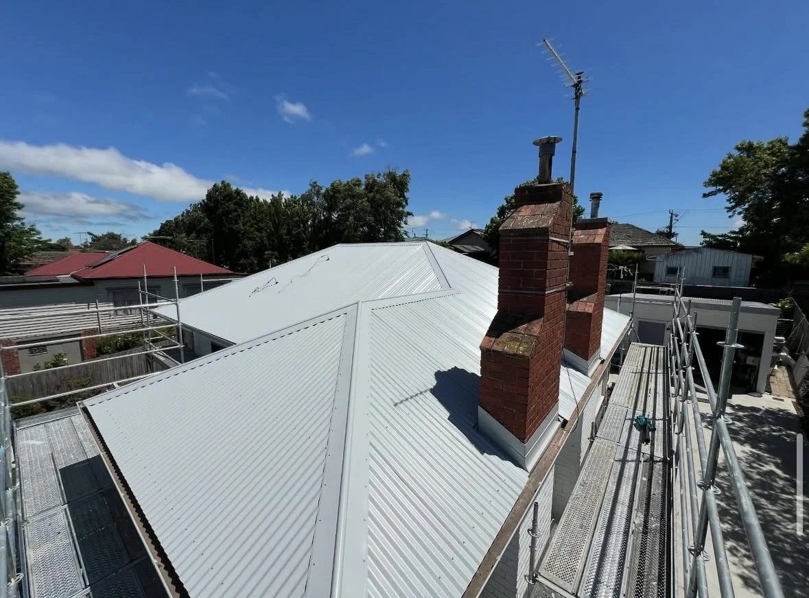 View of a house roof with white metal roofing, brick chimneys, a blue sky, and surrounding houses with scaffolding.