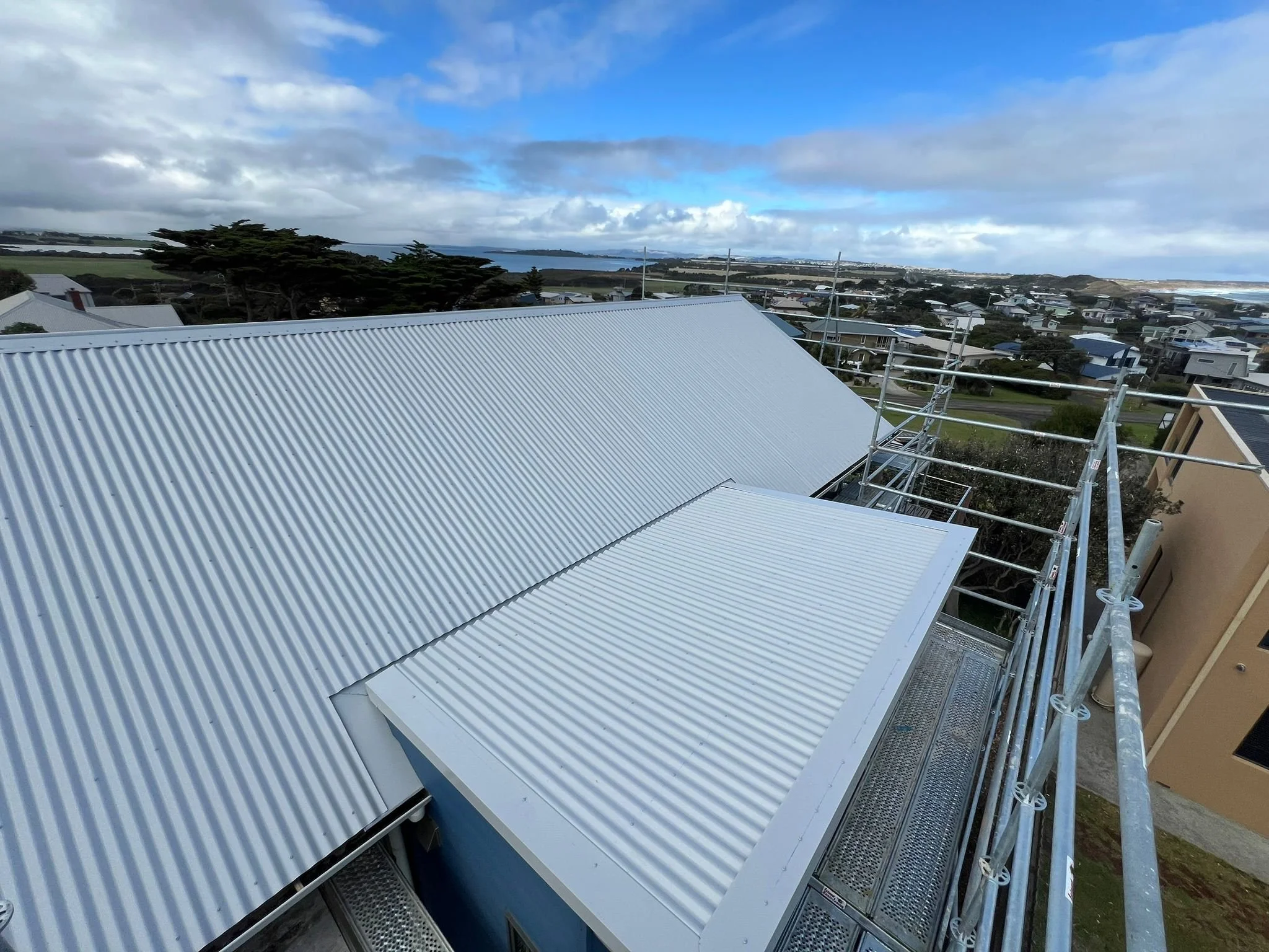 View of a corrugated metal roof on a building with a scaffolding structure on the side, overlooking a suburban neighborhood with numerous houses, trees, and a body of water in the distance under a cloudy sky.