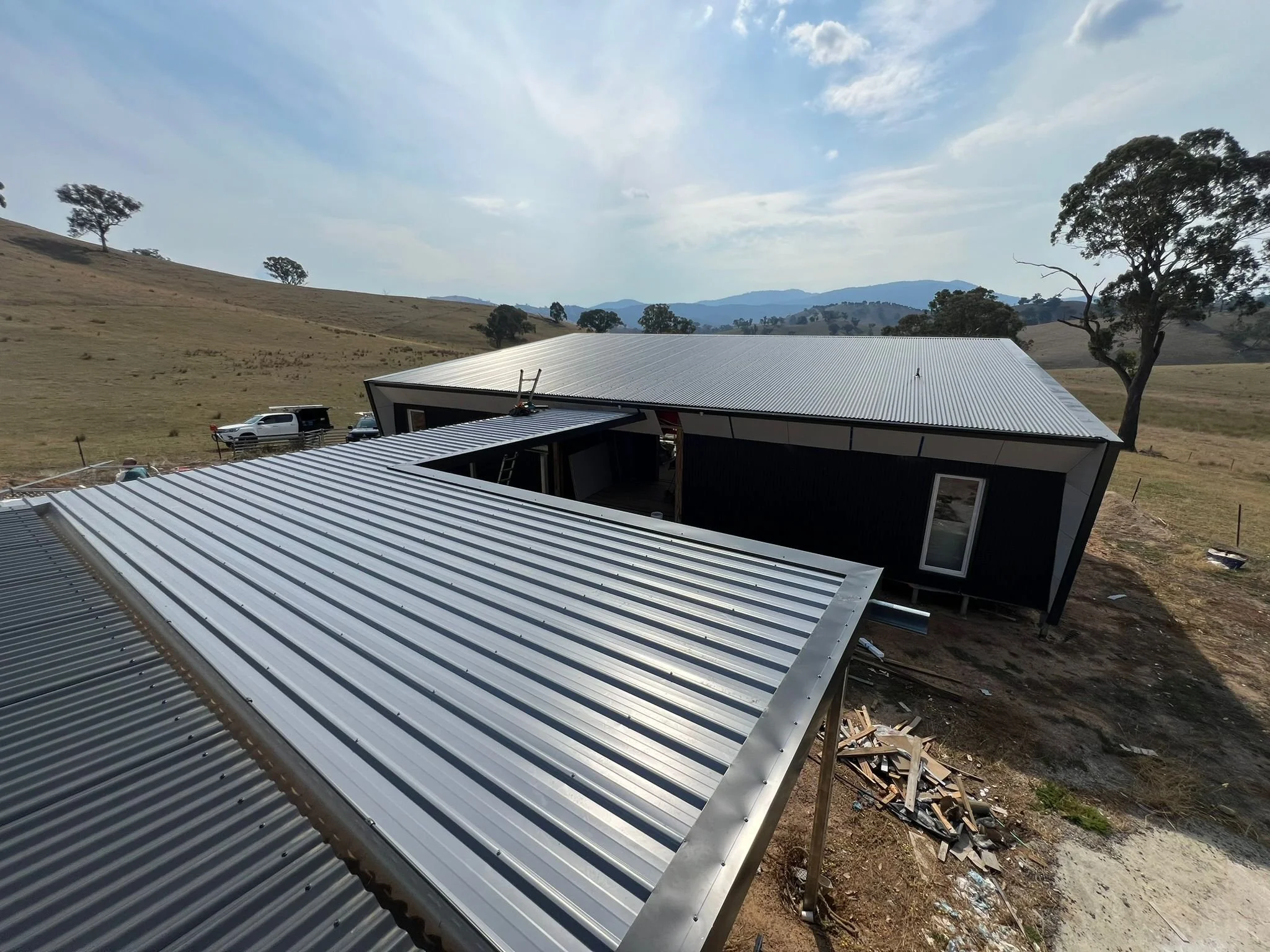 A building under construction with metal roofing panels in a rural area with hills, trees, and a partly cloudy sky in the background.