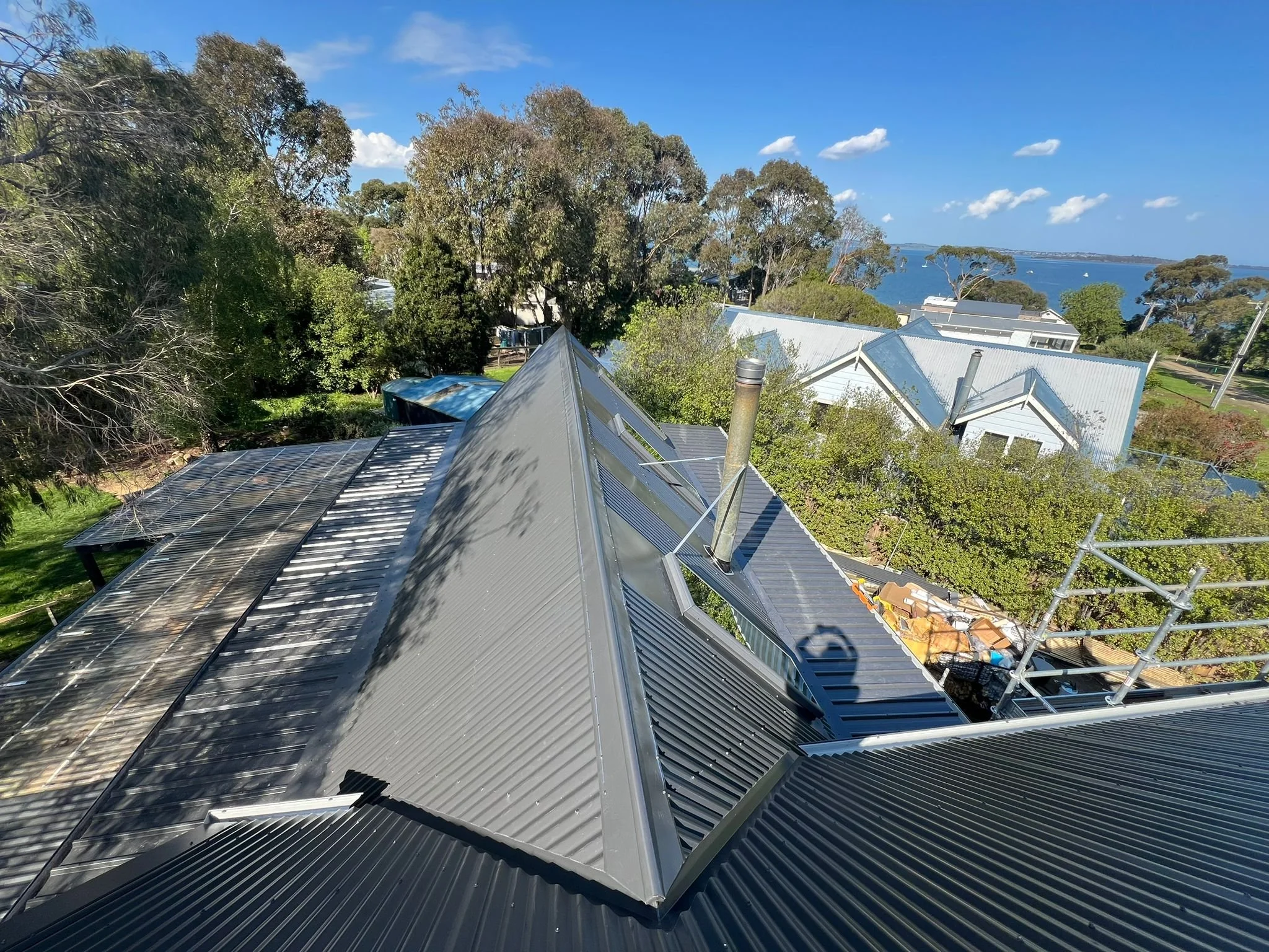 Aerial view of rooftops with solar panels and metal roofing, overlooking trees, houses, and a body of water in the distance under a blue sky with scattered clouds.