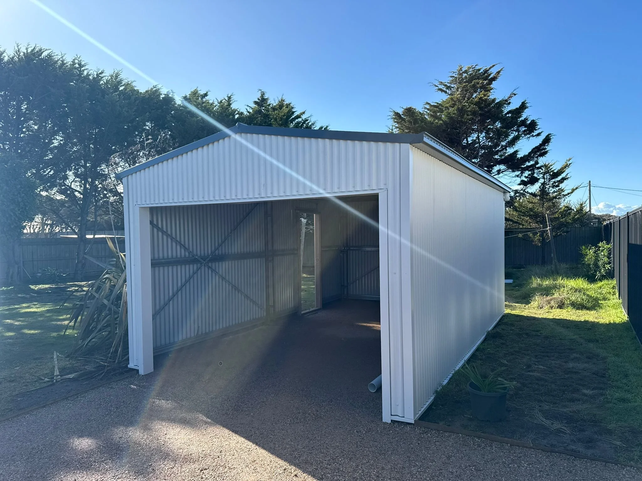 Empty metal shed in a backyard with trees and a fence, under a clear blue sky.