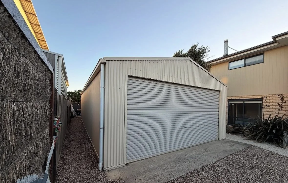 A beige metal garage with a roll-up door, located between two residential buildings, with a gravel and concrete driveway in front, during daytime.