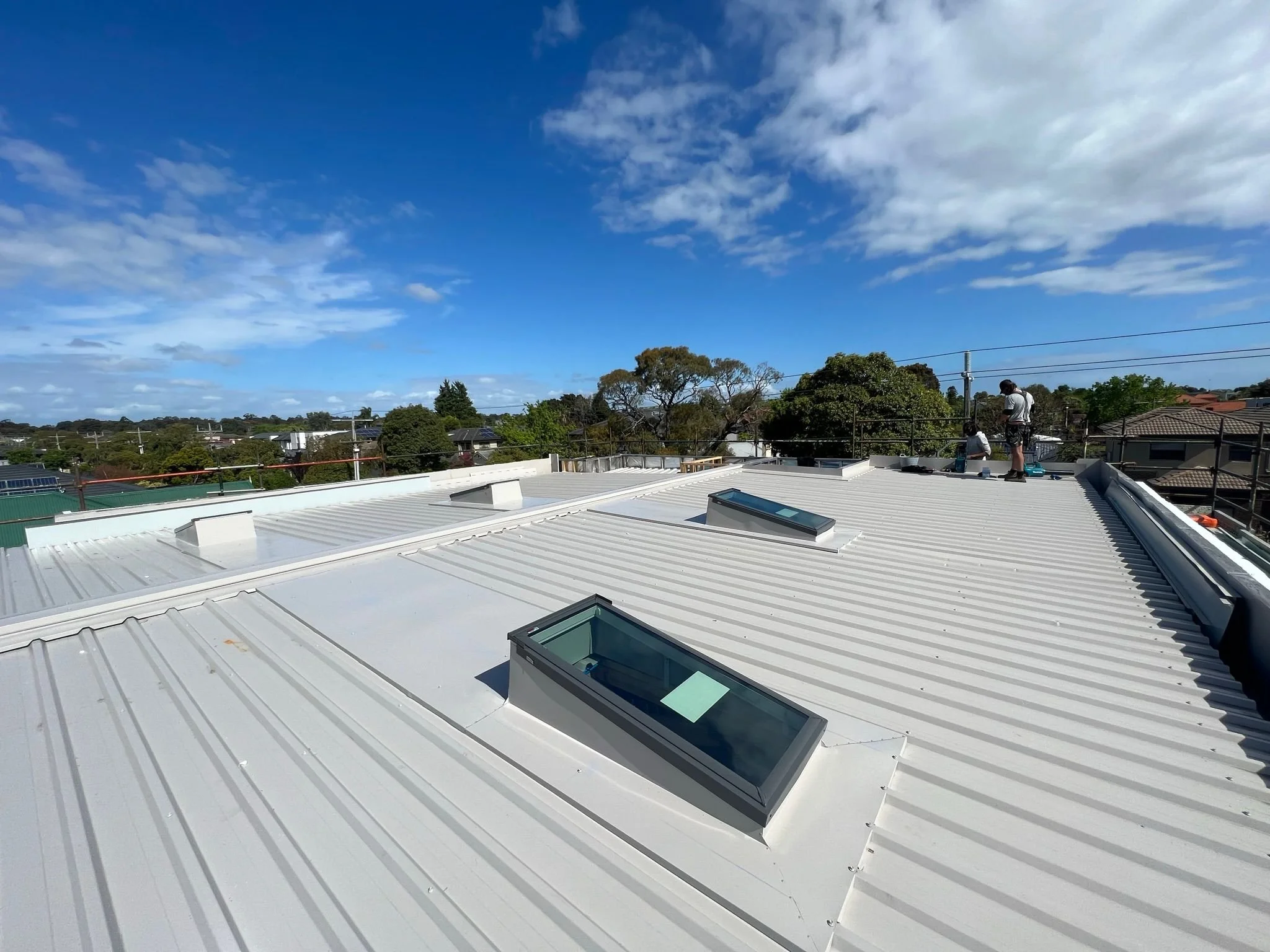 View of a white metal rooftop with skylights, under a blue sky with scattered clouds. Two workers are on the roof, near the edge, working on safety railings.