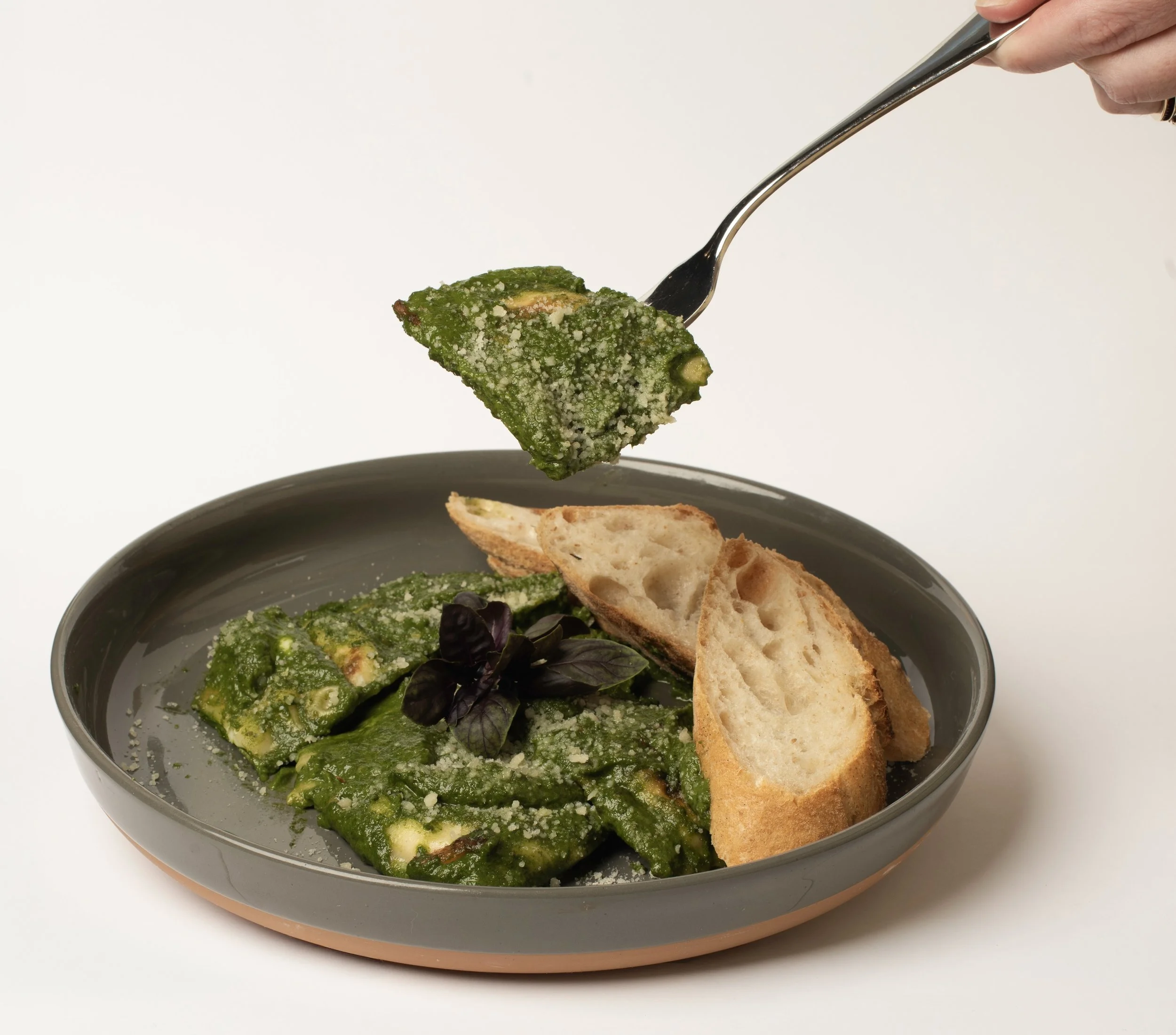 A hand holds a fork with a piece of green pesto bread over a bowl of salad that contains leafy greens, avocado slices, and black olives, with additional slices of bread in the bowl.