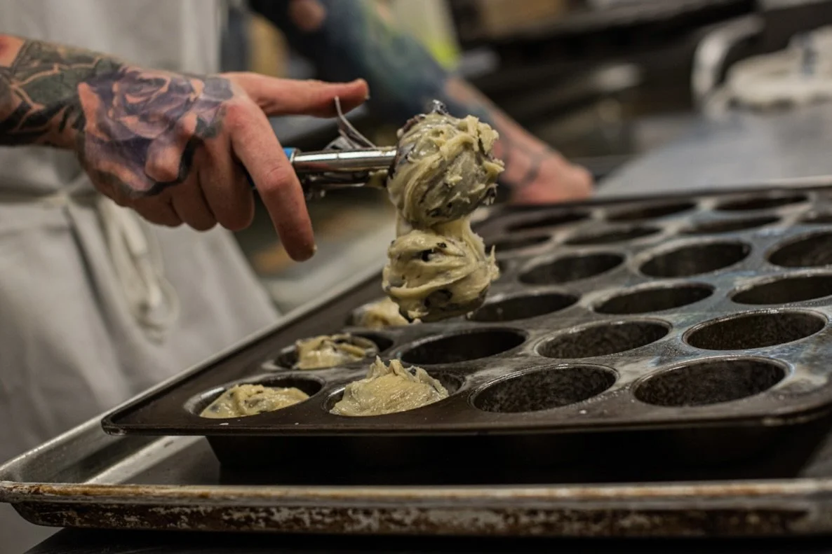 A person with tattoos on their hand is scooping dough into a metal baking tray with multiple circular molds, likely for making muffins or cupcakes, in a kitchen.