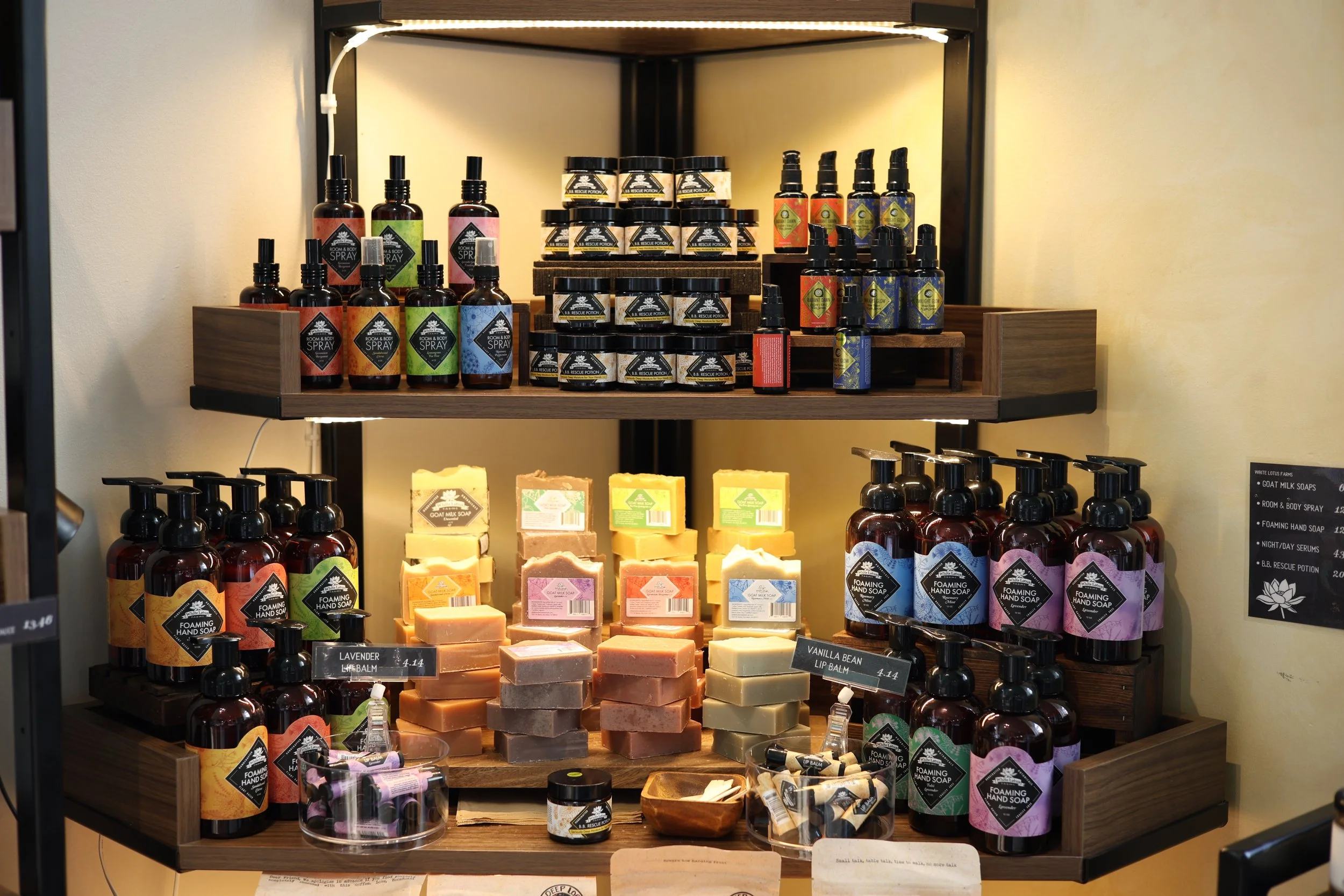Display of various soap and lotion bottles and bars arranged on wooden shelves, with labels indicating scents and types, in a retail store setting.