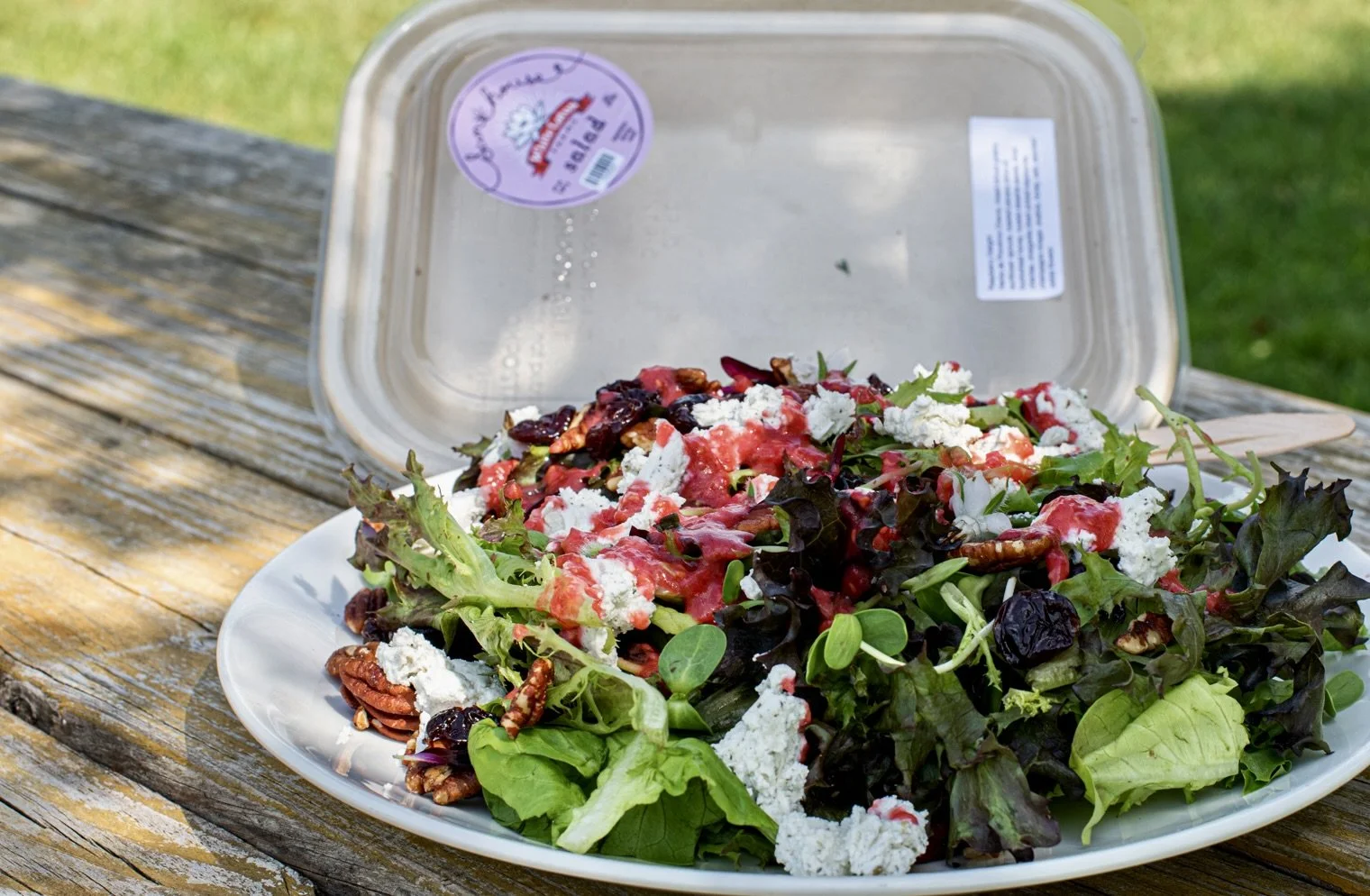 A plate of mixed greens salad with nuts, crumbled cheese, and berry vinaigrette, with a plastic food container in the background on a wooden outdoor table.