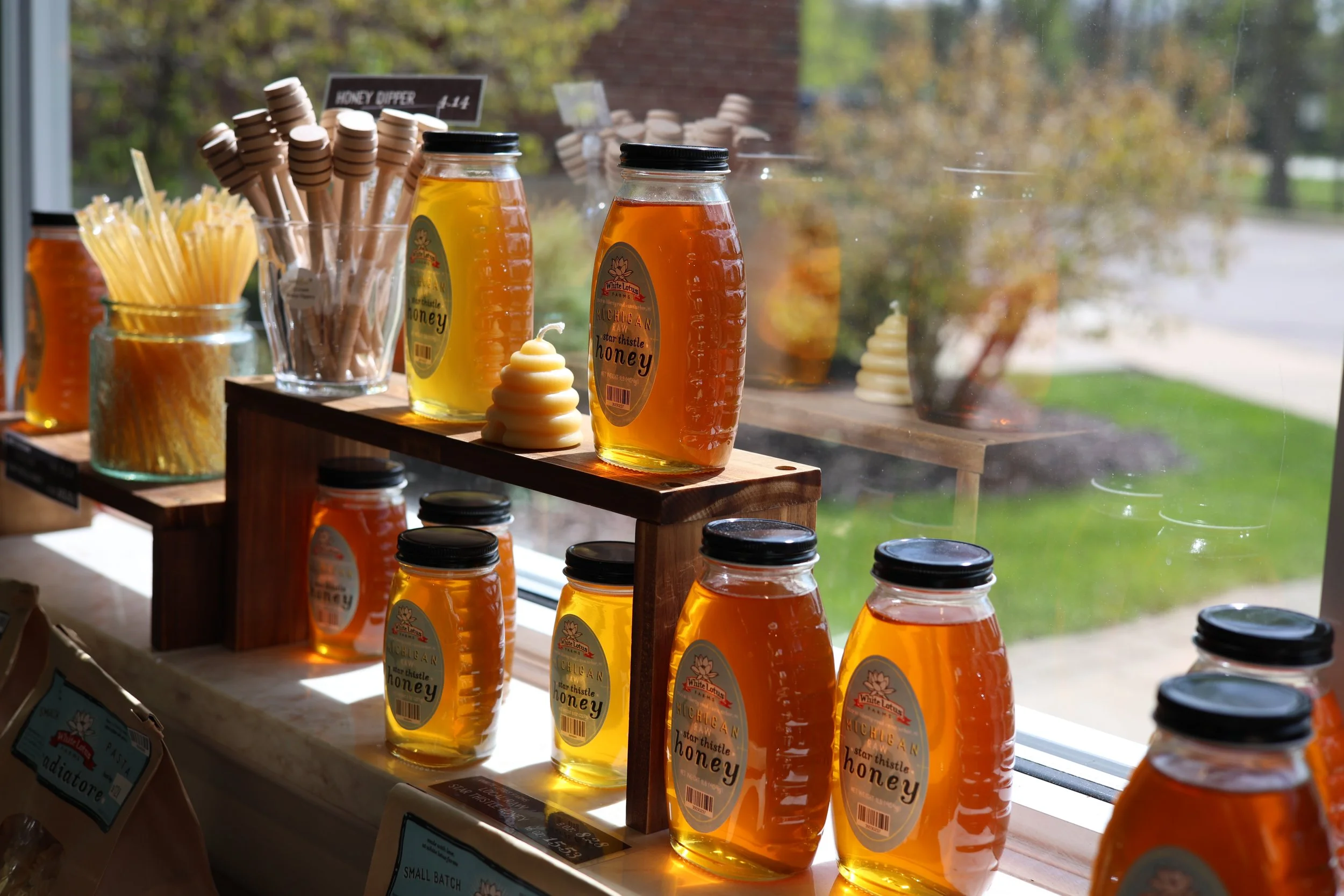 Several jars of honey on wooden shelves near a window with a view of trees and grass outside, along with honey sticks and a honey dipper in jars.
