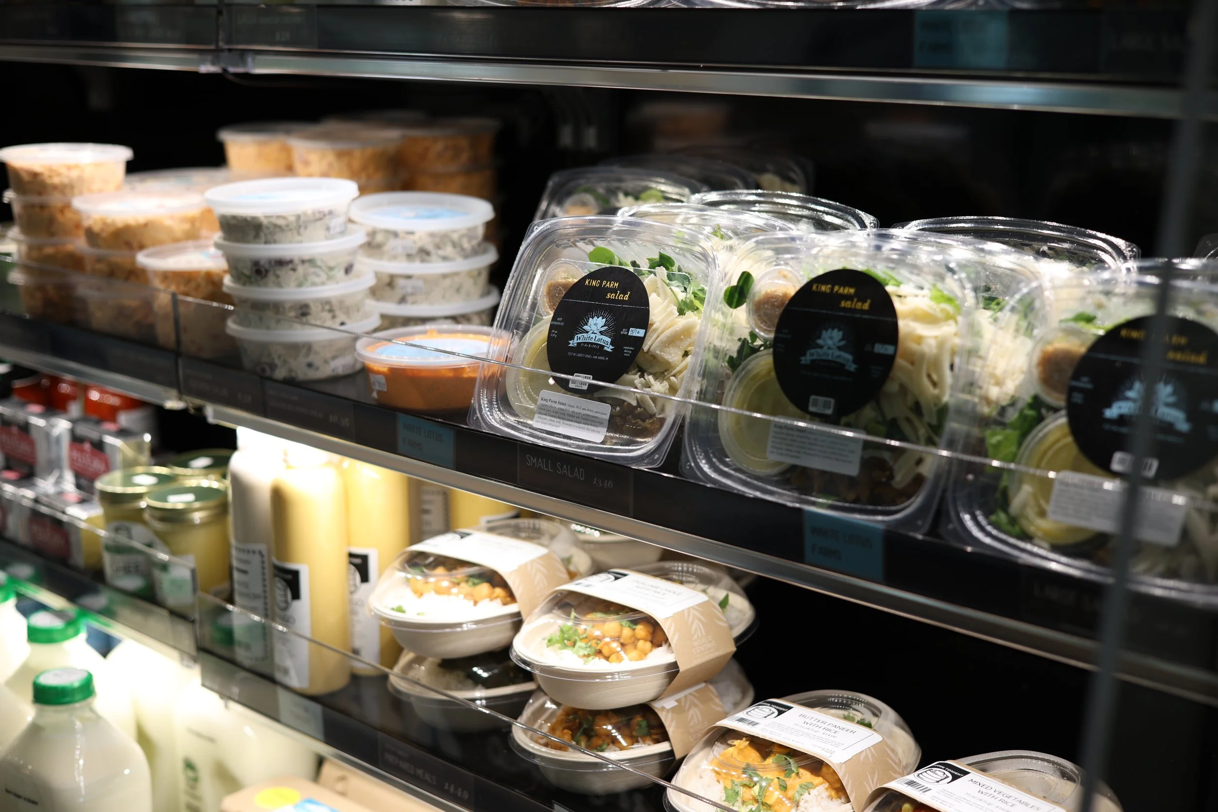 Display of pre-packaged salads in plastic containers on supermarket shelves, including mixed greens, spinach, and pasta salads.