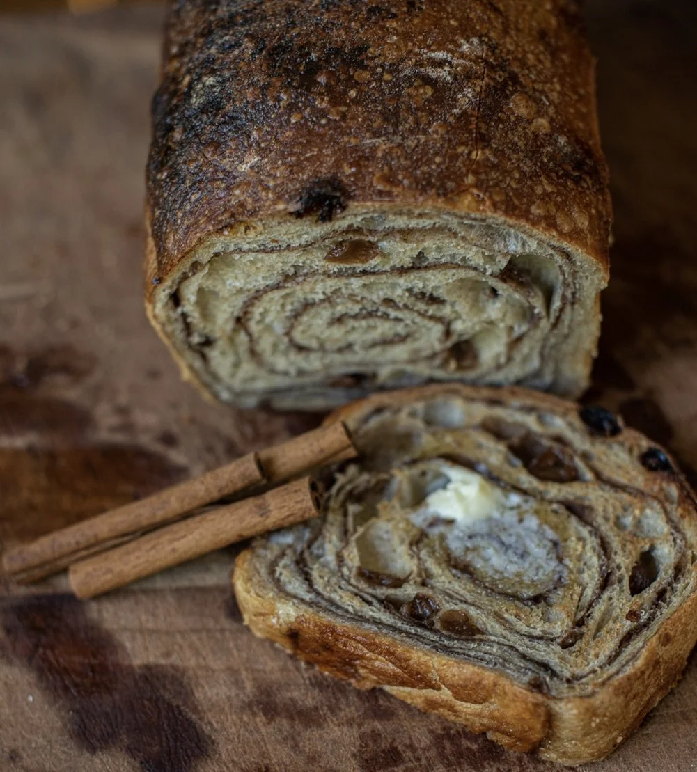Close-up of a cinnamon roll with swirled layers, topped with sugar and cinnamon, placed on a wooden surface with cinnamon sticks nearby.