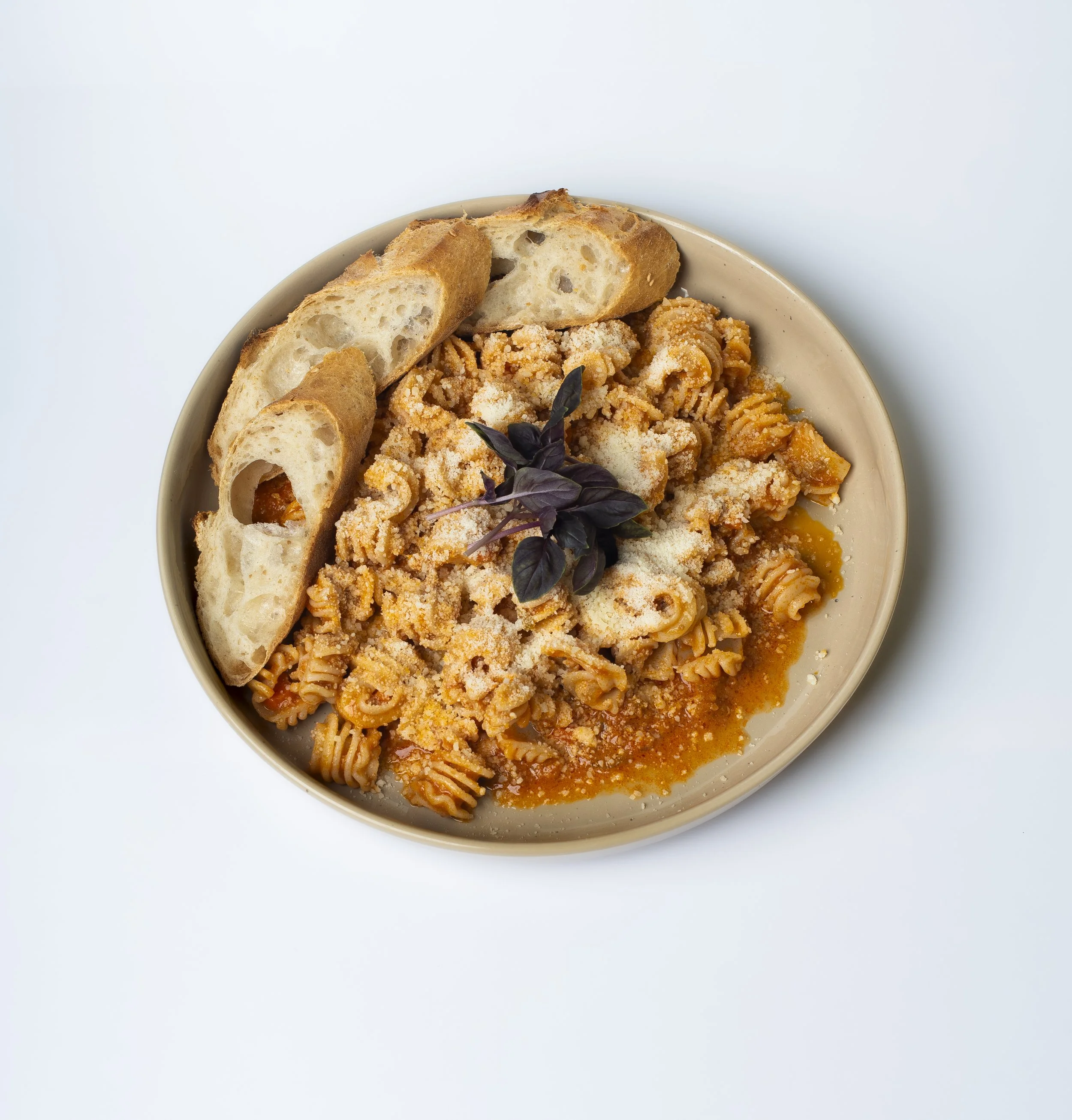 A beige bowl containing spaghetti with tomato sauce, grated cheese, and garnished with purple basil leaves, with slices of rustic bread on the side, against a white background.