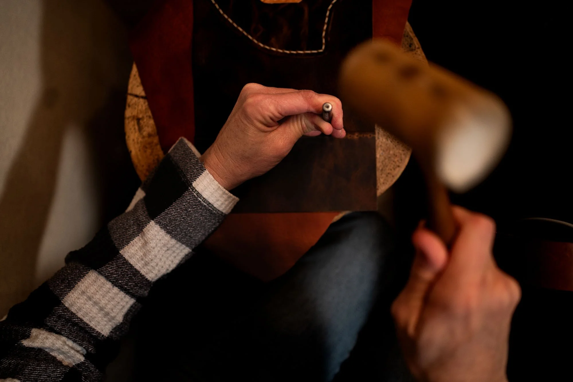 A person working with a piece of brown leather, sewing or crafting, using a tool in their right hand, while their left hand holds the leather steady. They are wearing a plaid shirt with black and white checks.