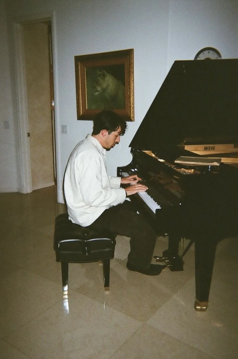 A man in a white shirt playing a black grand piano in a room with beige tile flooring, a landscape painting of a wolf on the wall, and a clock above the piano.