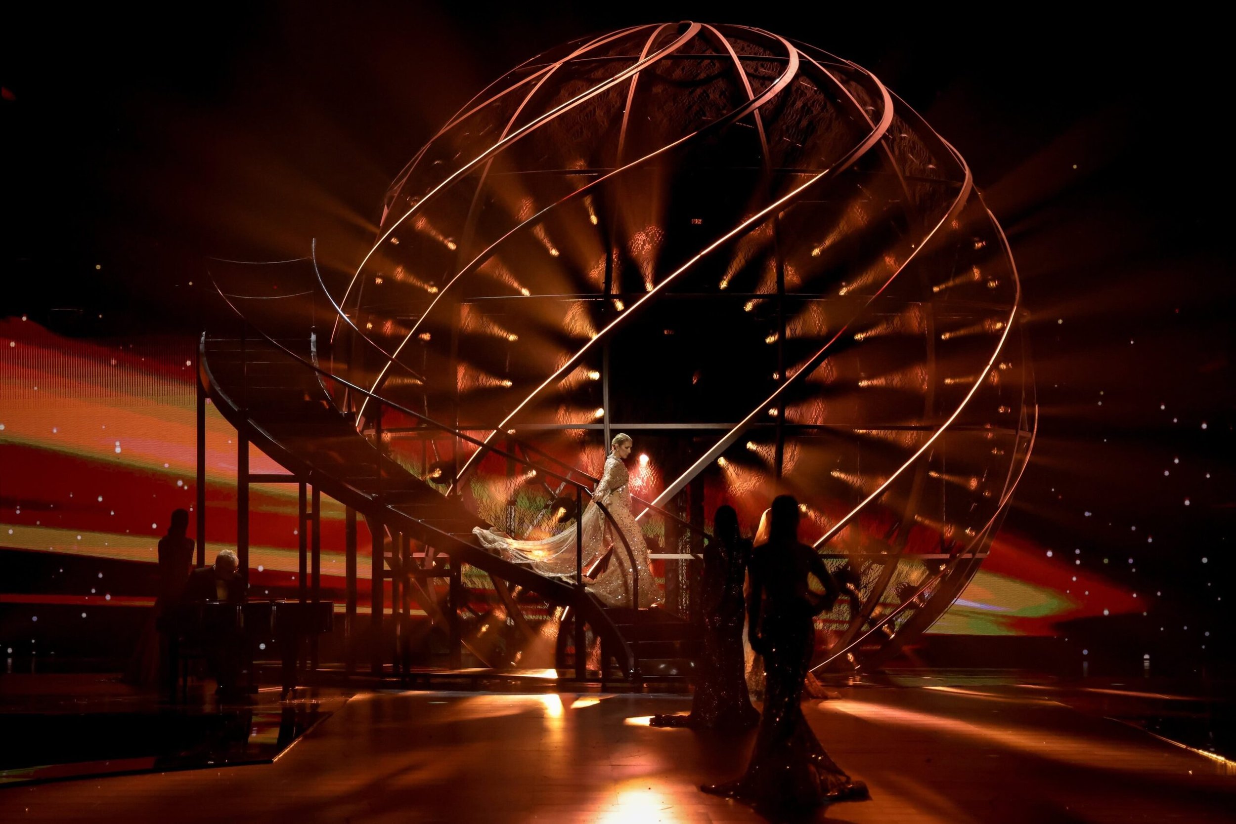 A stage with a large, illuminated, spiral, spherical structure, gold lighting, and people dressed in formal or glamorous attire, with a person walking down the stairs and a woman in a gown at the center.