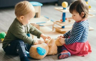Two young children playing together with a teddy bear on a playroom floor.