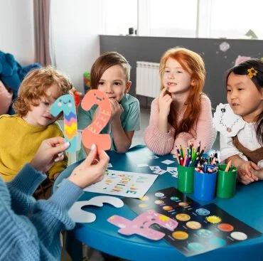 Children sitting around a blue table engaging in a learning activity with colorful paper cutouts and art supplies.