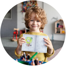 A young child with curly red hair holding an open drawing book with colorful drawings, smiling in a cozy room with toys and decorations in the background.