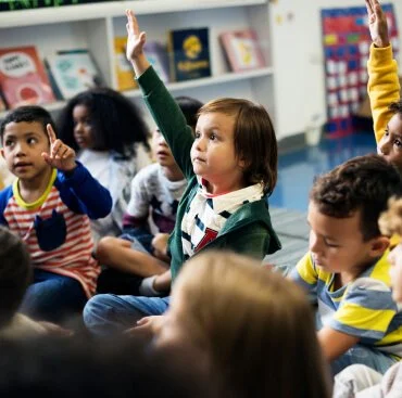 Young children in a classroom, with some raising their hands, participating in a learning activity.