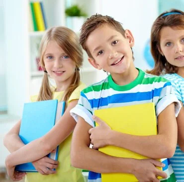 Three children standing indoors, holding books and smiling