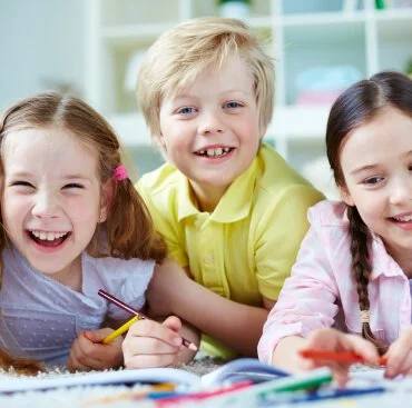 Three children smiling and drawing at a table in a classroom.