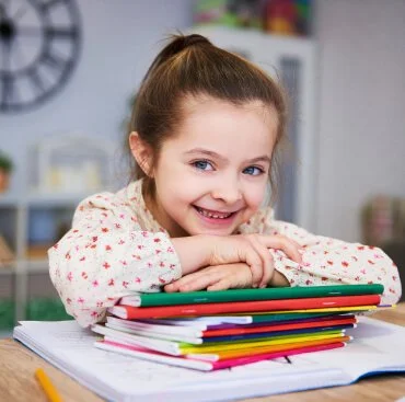 A young girl with a ponytail and a floral blouse smiling at the camera, resting her chin on her folded arms on a stack of colorful notebooks and papers at a desk in a classroom or study room.