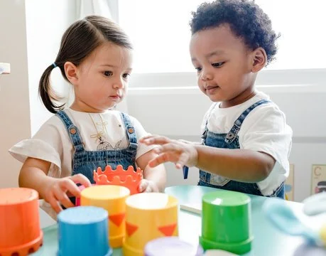 Two young children engaging in a play activity with colorful stacking cups and toys at a table.