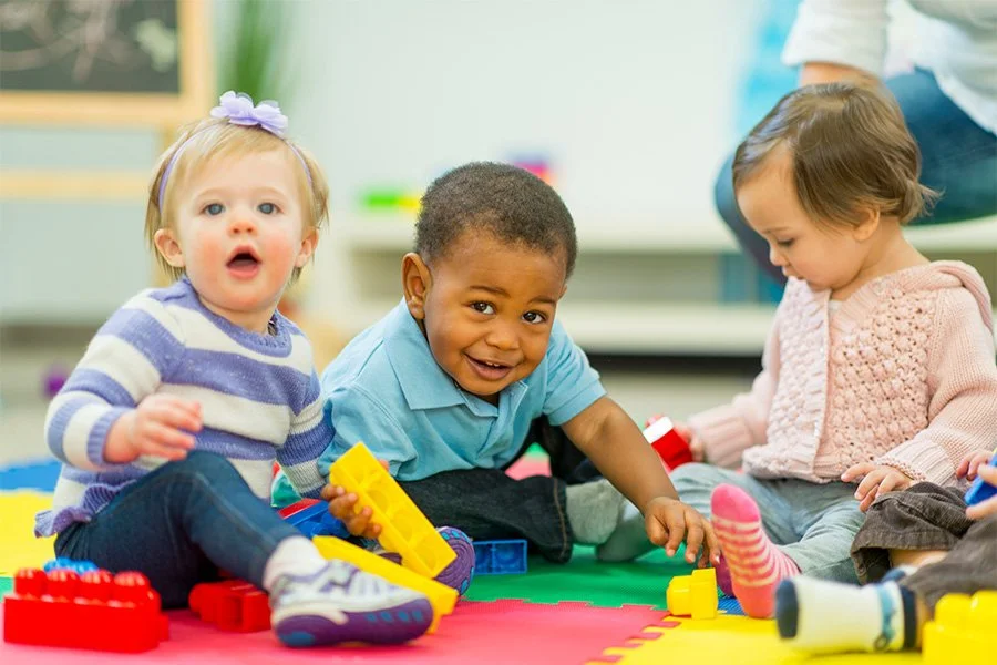 Three young children playing with colorful building blocks on a foam mat in a classroom or daycare setting.