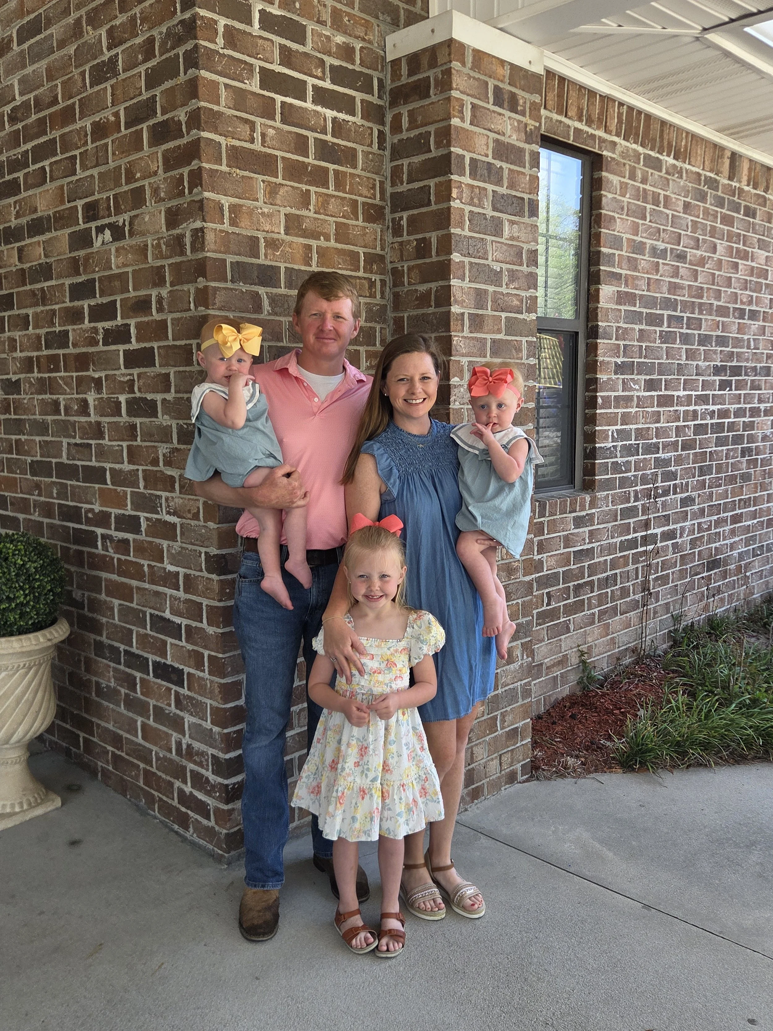 A family of five standing outside near a brick building with a large window. The father, wearing a pink shirt, is holding two young girls with bows in their hair. The mother, wearing a blue dress, is holding another young girl. There is a young girl standing in front with a floral dress. They are all smiling.