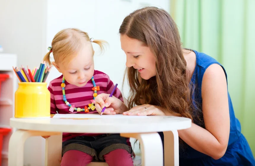 A woman and a young girl sitting at a white table, coloring together with colored pencils in a classroom or playroom with green curtains in the background.