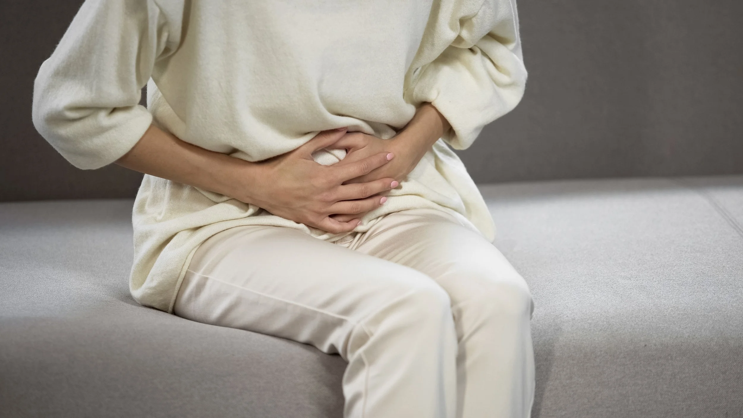 A person sitting on a gray sofa, wearing a cream-colored, loose-fitting outfit, clutching their stomach with both hands, indicating stomach pain or discomfort.