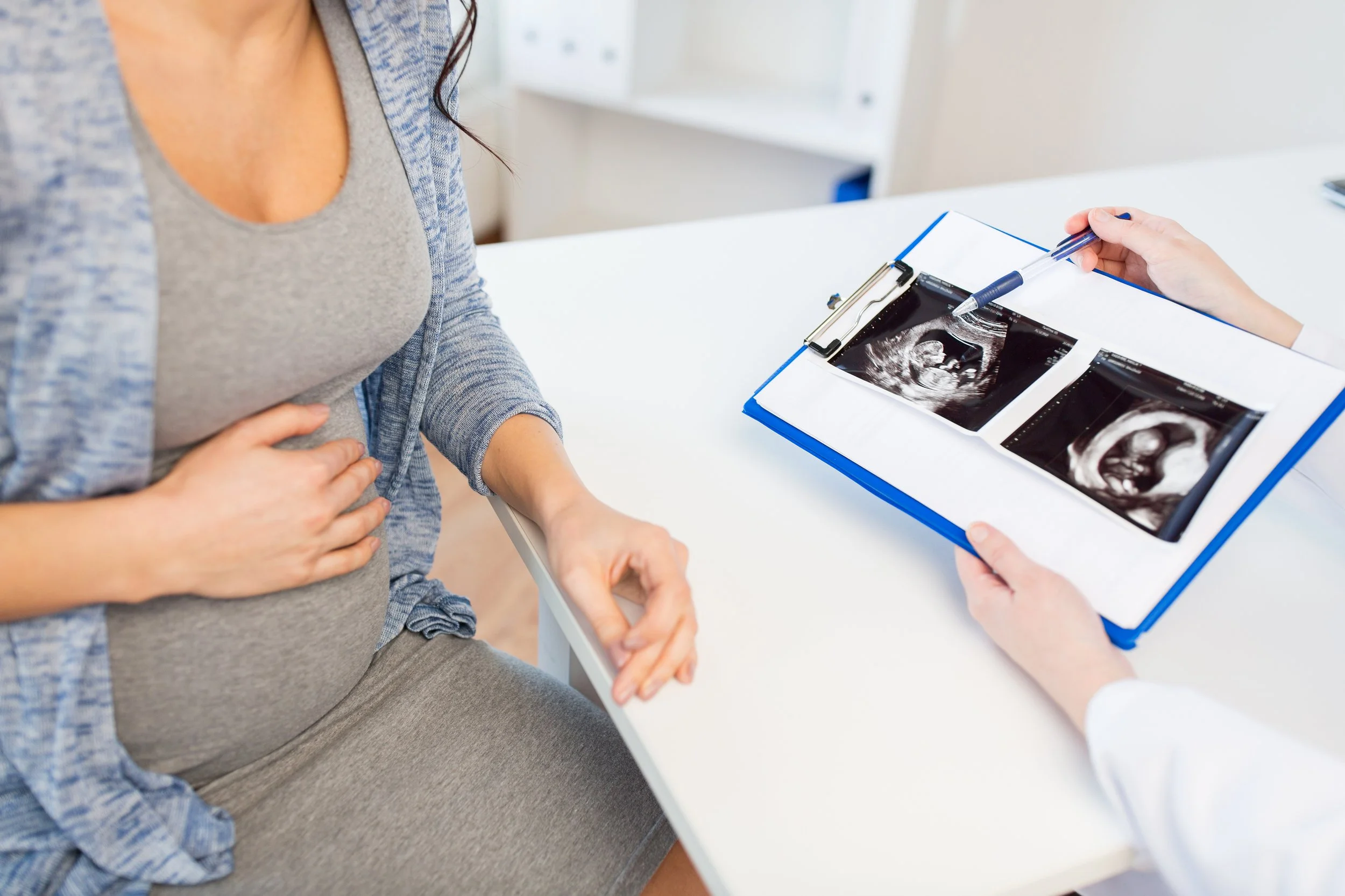 A pregnant woman sitting at a table with her hands resting on her belly while a healthcare professional shows her ultrasound images.