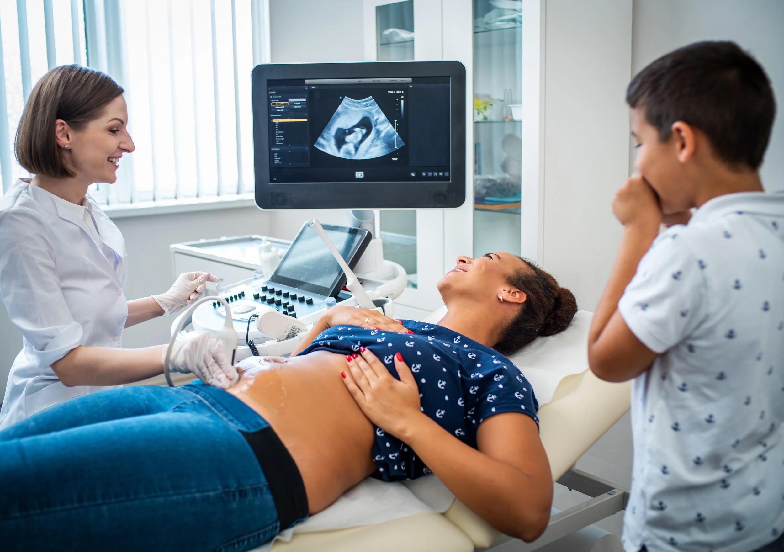 A pregnant woman lying on an examination table during an ultrasound, with a healthcare professional and a young man present in the room.