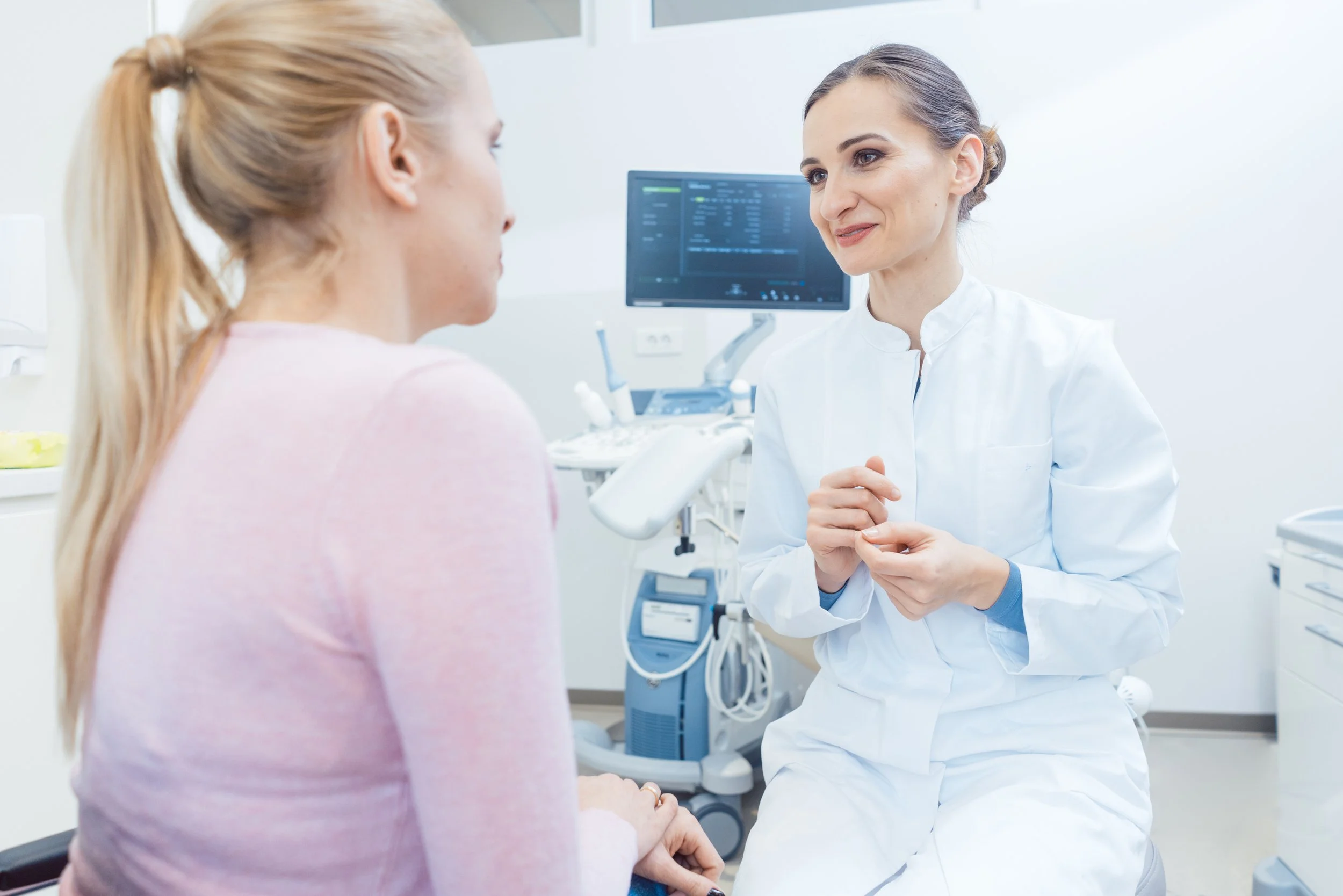 A woman with blonde hair in a ponytail sitting in a medical examination room, talking to a healthcare professional dressed in white, with medical equipment and a monitor showing health data in the background.