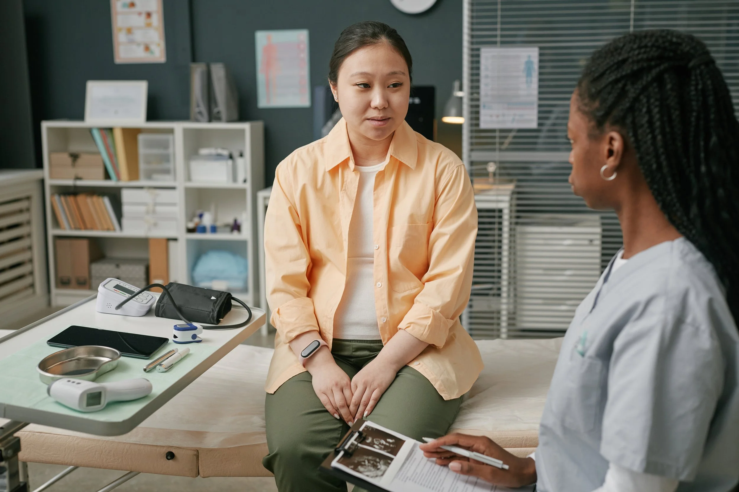 A healthcare setting with a woman in a peach shirt sitting on an examination table, talking to a nurse or doctor holding medical images or reports. Medical equipment and supplies are on a nearby tray.